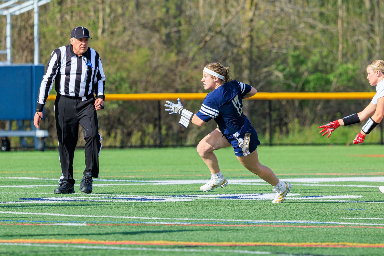 A lacrosse player in navy uniform sprints on turf field while a referee in stripes observes during an outdoor game.