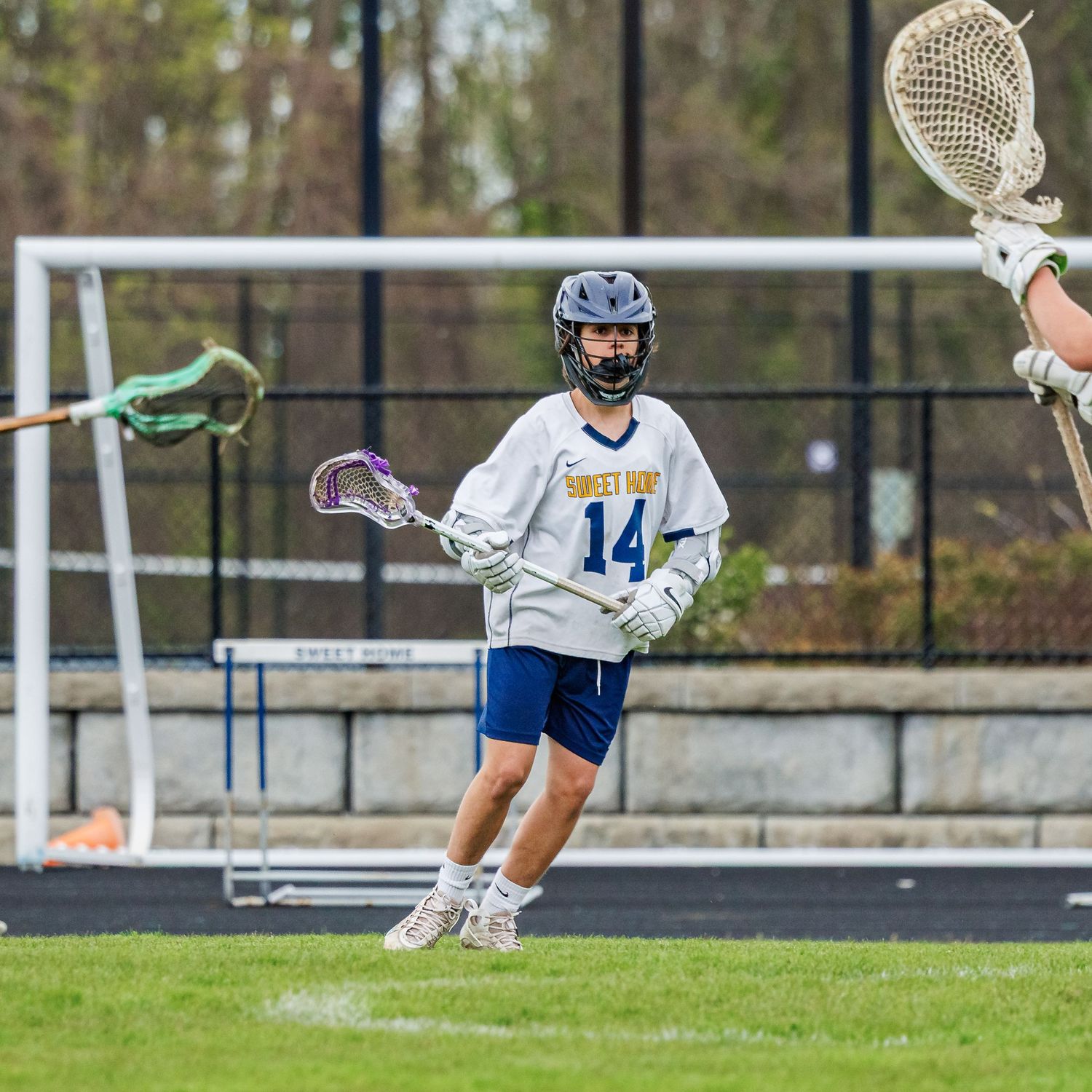Lacrosse player wearing jersey number 14 in white and blue uniform holds stick on field near goal.