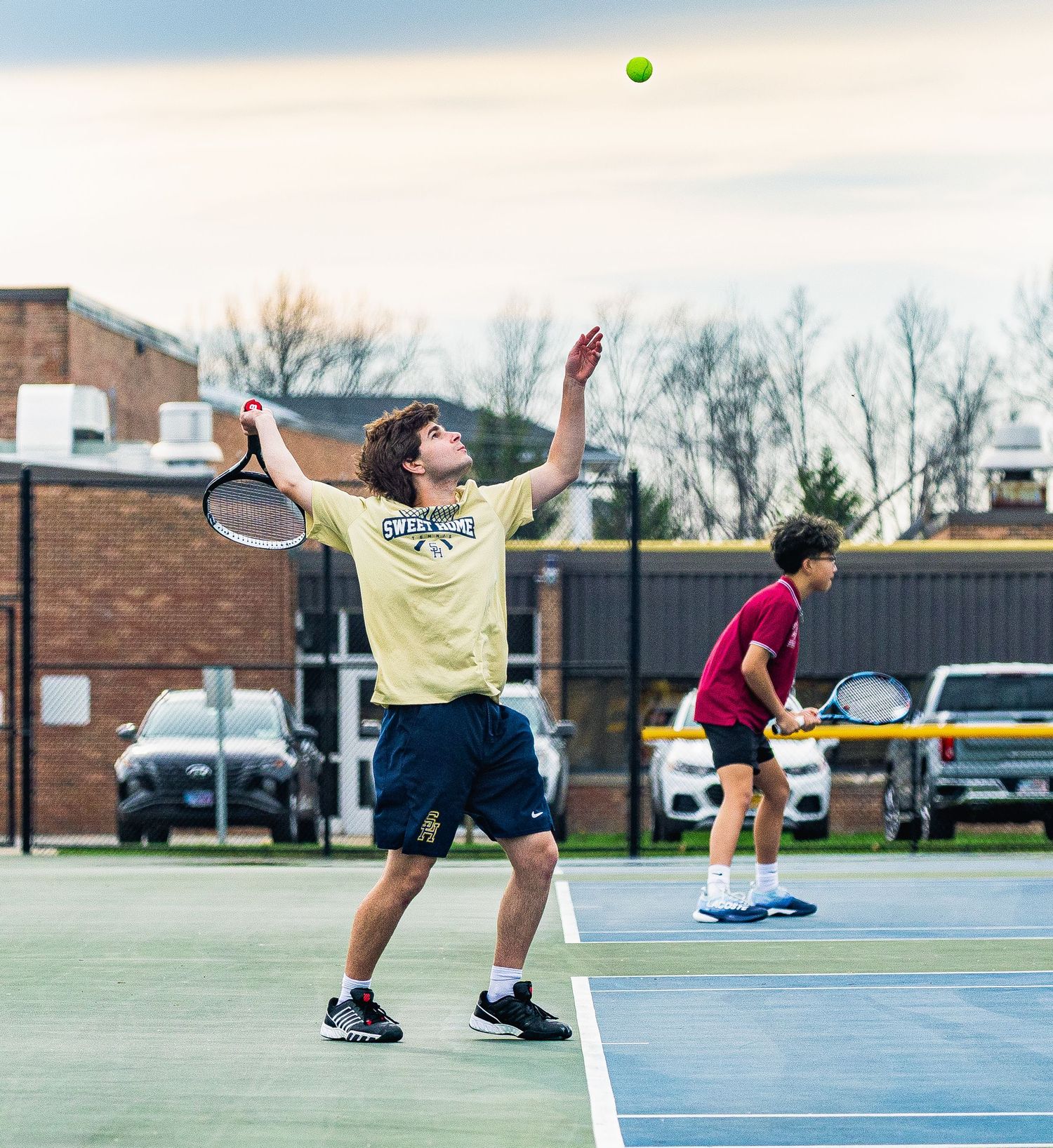 Two tennis players practice on an outdoor court, one serving the ball high into the air in a yellow shirt.