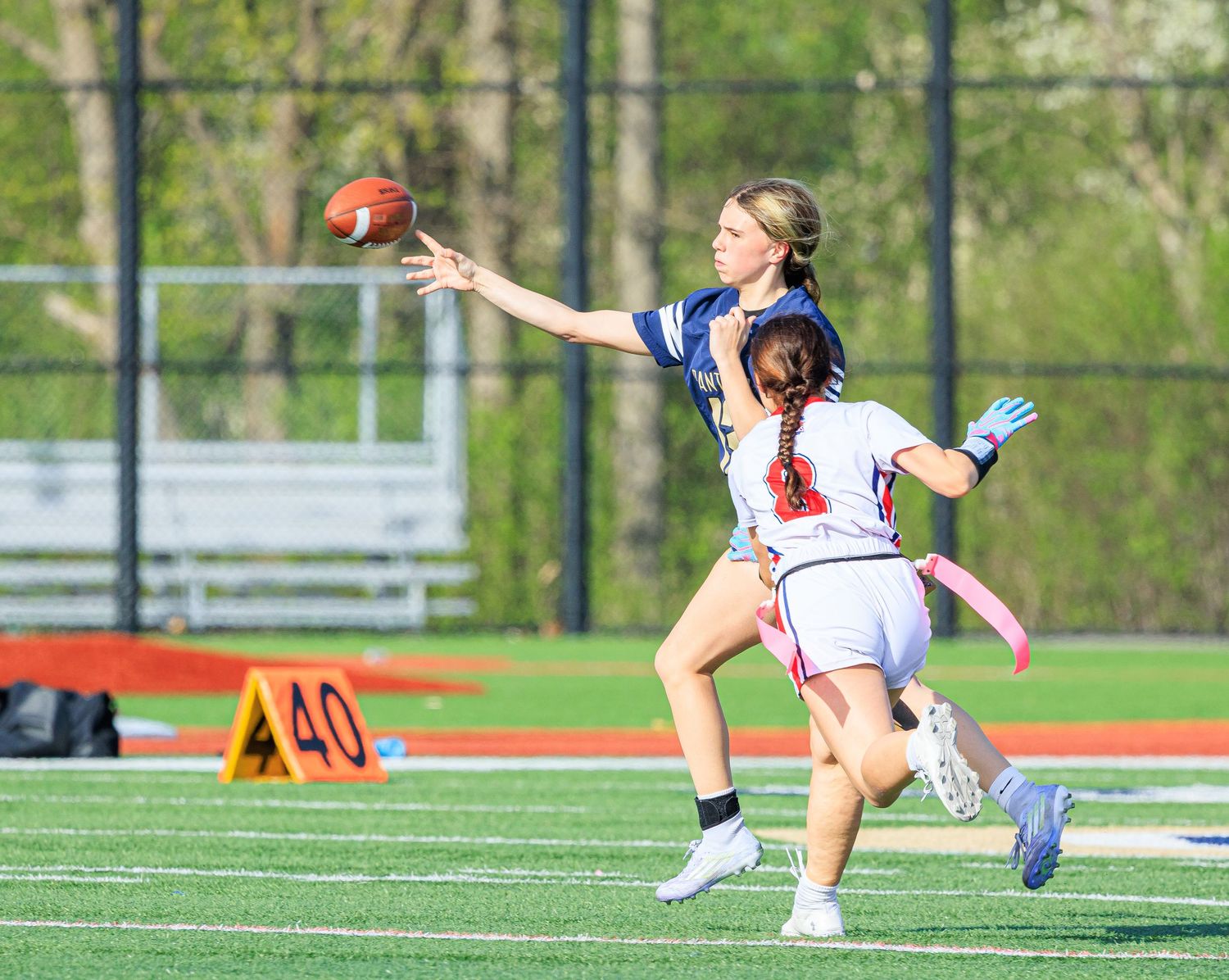Female flag football player reaches to catch pass while defender pressures her.