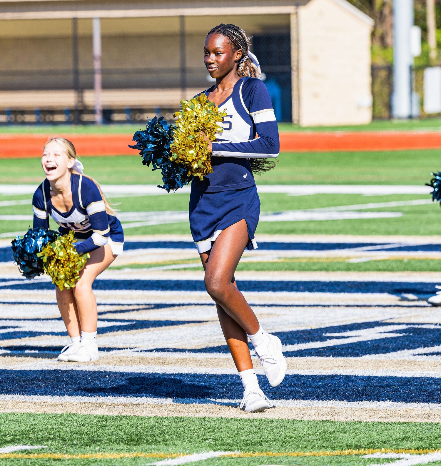 Cheerleader in blue uniform runs with gold pom-poms on a football field.