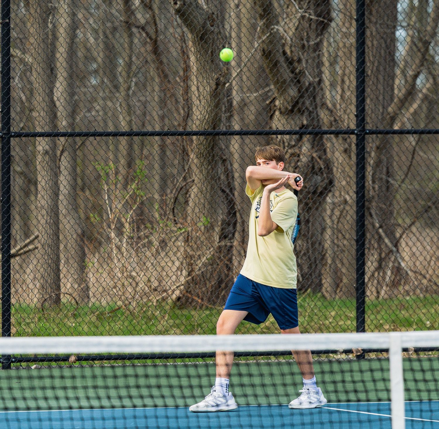 A young tennis player in a yellow shirt and blue shorts hits a ball on an outdoor court backed by a black fence.