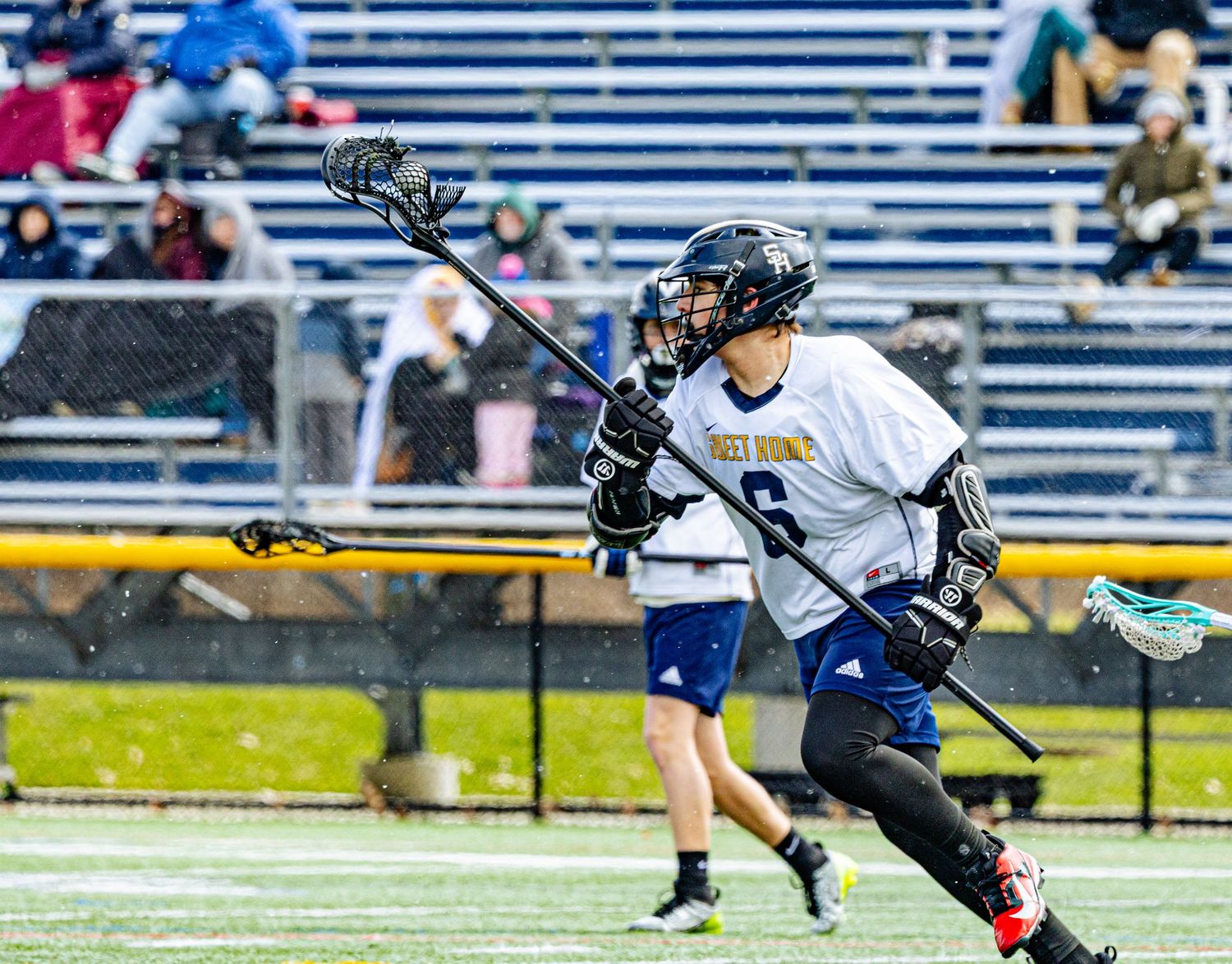 Lacrosse player #6 in blue and white uniform cradling the ball during an outdoor match.