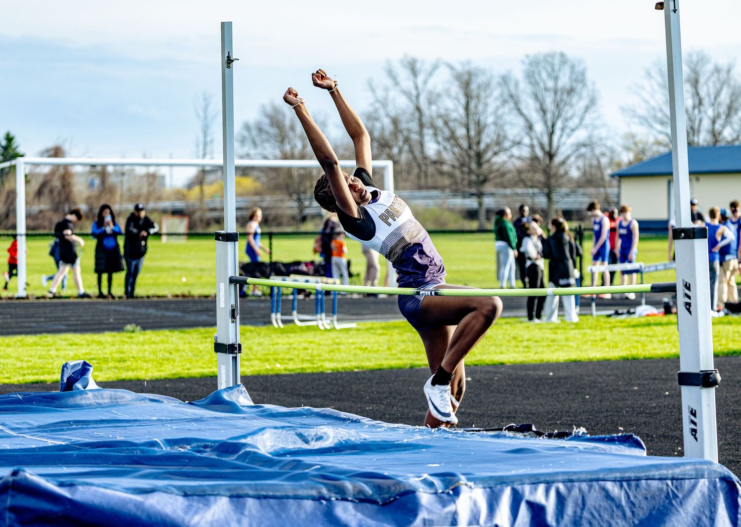 Athlete performs high jump at outdoor track meet, soaring over the bar with arms raised against a cloudy sky.