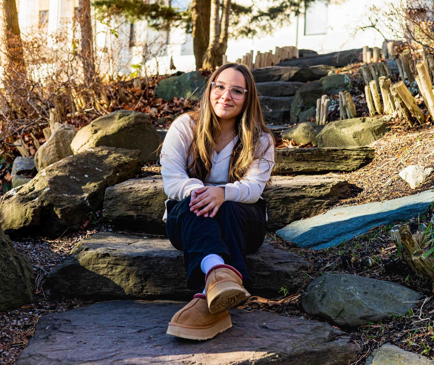 Someone in casual winter attire sits on large rocks outdoors during golden hour lighting.