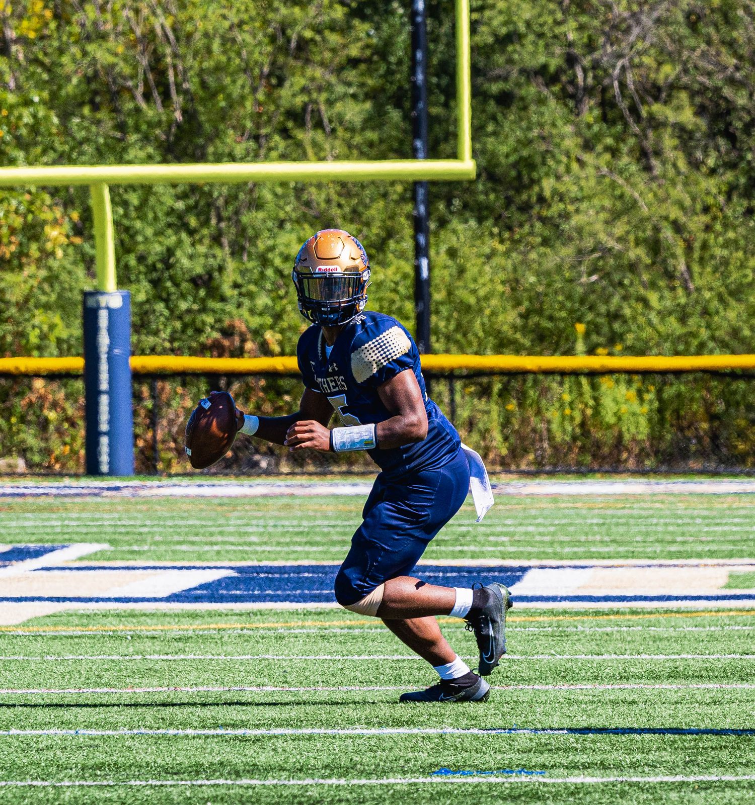 College football quarterback in blue and gold uniform drops back to pass on green turf field with yellow goalpost behind.