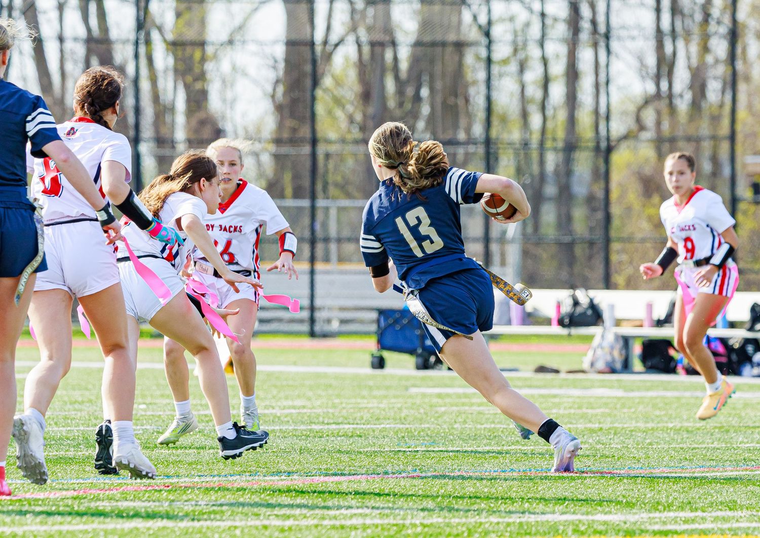 Female lacrosse player #13 in navy uniform sprints past defenders during a competitive outdoor game on a green turf field.