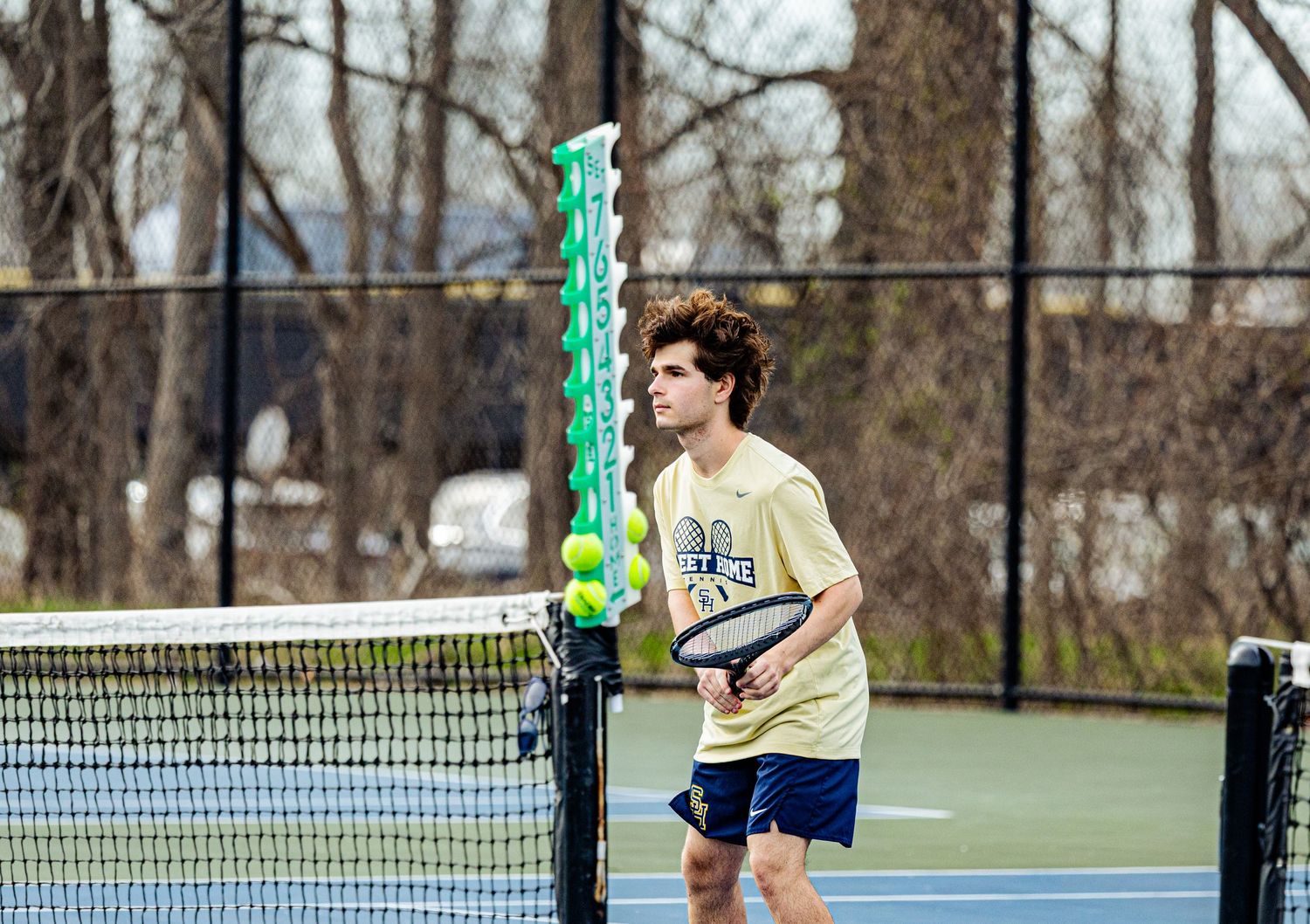 A young male tennis player holds a racket near the net on an outdoor court with tennis balls on a ball holder nearby.