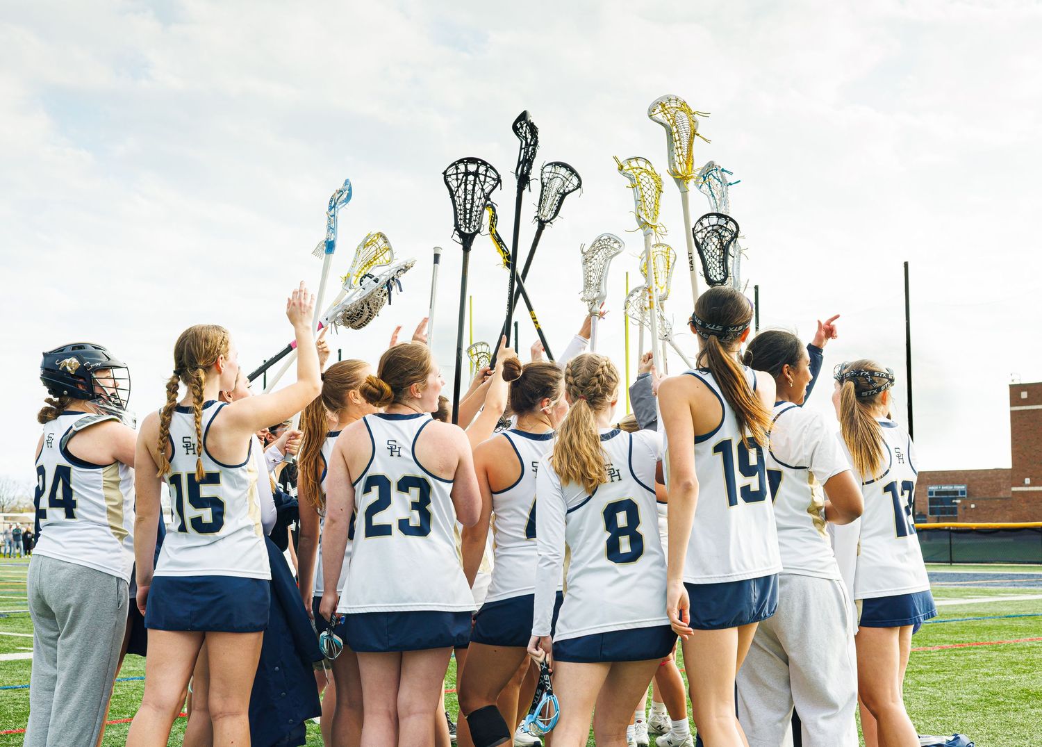 Girls lacrosse team huddles together raising sticks in team celebration wearing white uniforms on outdoor field.