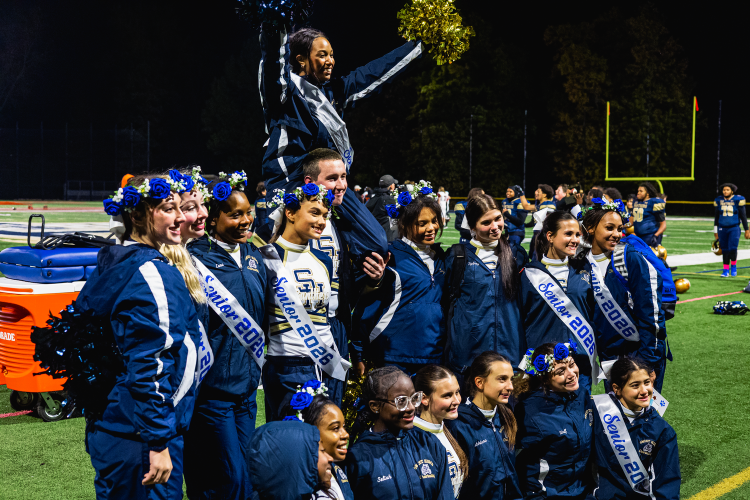 Cheerleaders in blue and gold uniforms pose together on a football field at night, with one member lifted in the air.