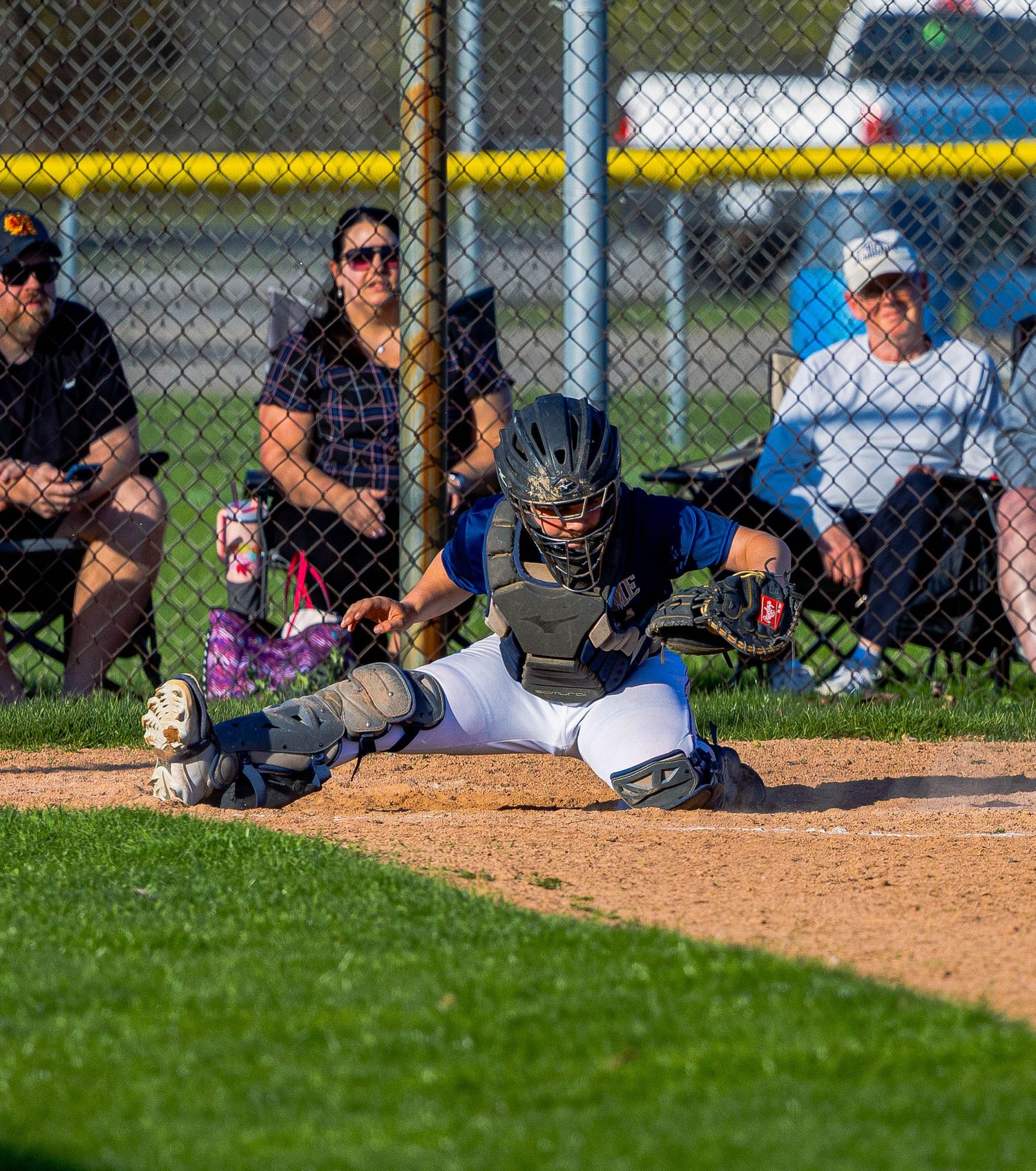 Baseball catcher in protective gear blocks home plate while fielding a play during youth baseball game.
