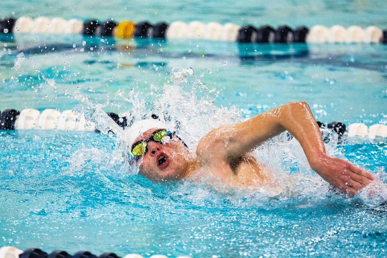 Competitive swimmer doing freestyle stroke in pool, wearing goggles and swim cap, splashing water during race.