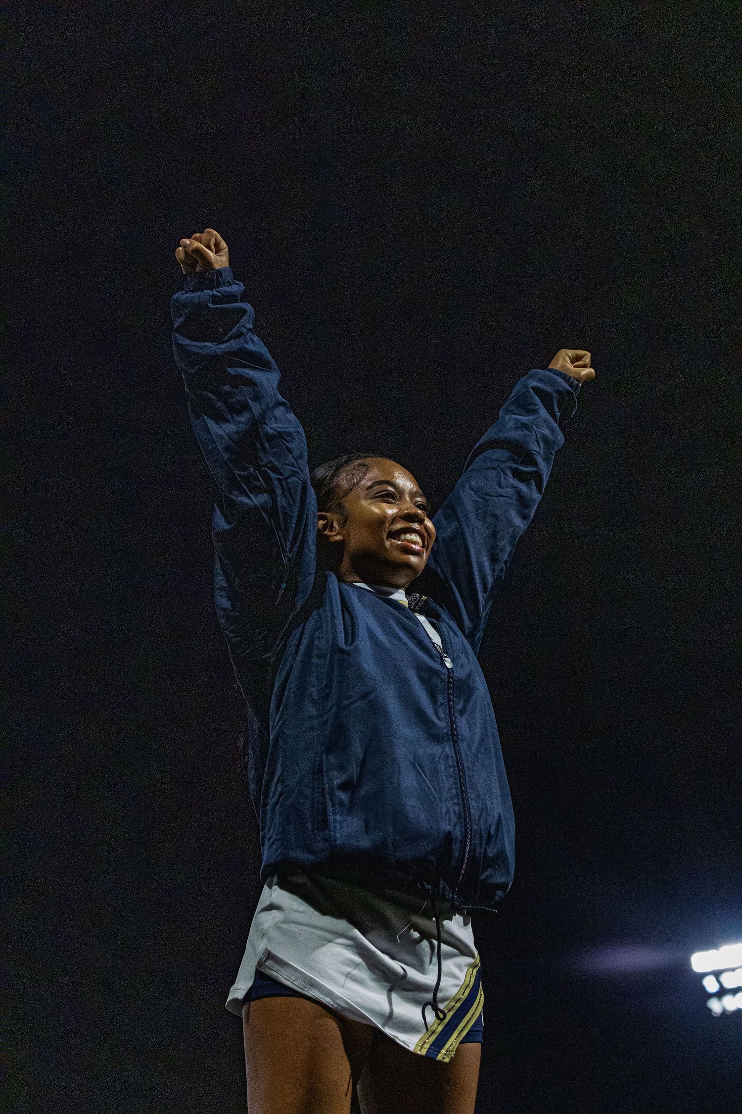 Young Black woman in blue jacket raises both arms in victory celebration at a nighttime sports event.