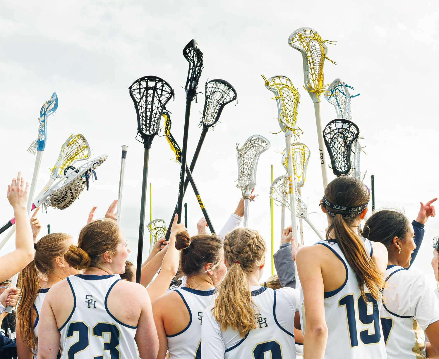 Female lacrosse players raise sticks skyward in team huddle wearing white and navy uniforms outdoors.