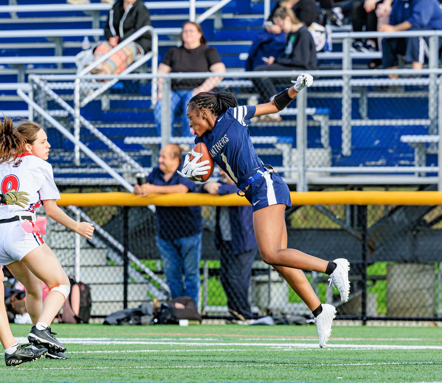 Female lacrosse player in blue uniform sprinting with ball during competitive game on turf field with spectators in bleachers.
