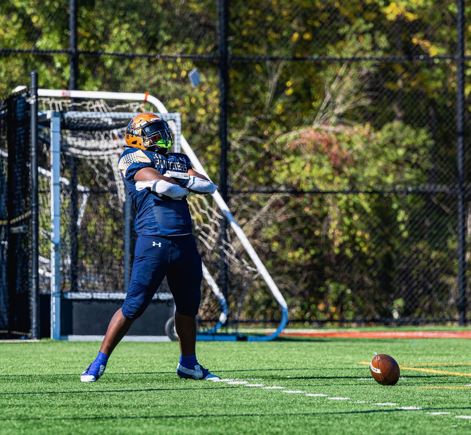 Football player in navy uniform winds up for a kick on an outdoor turf field during practice.
