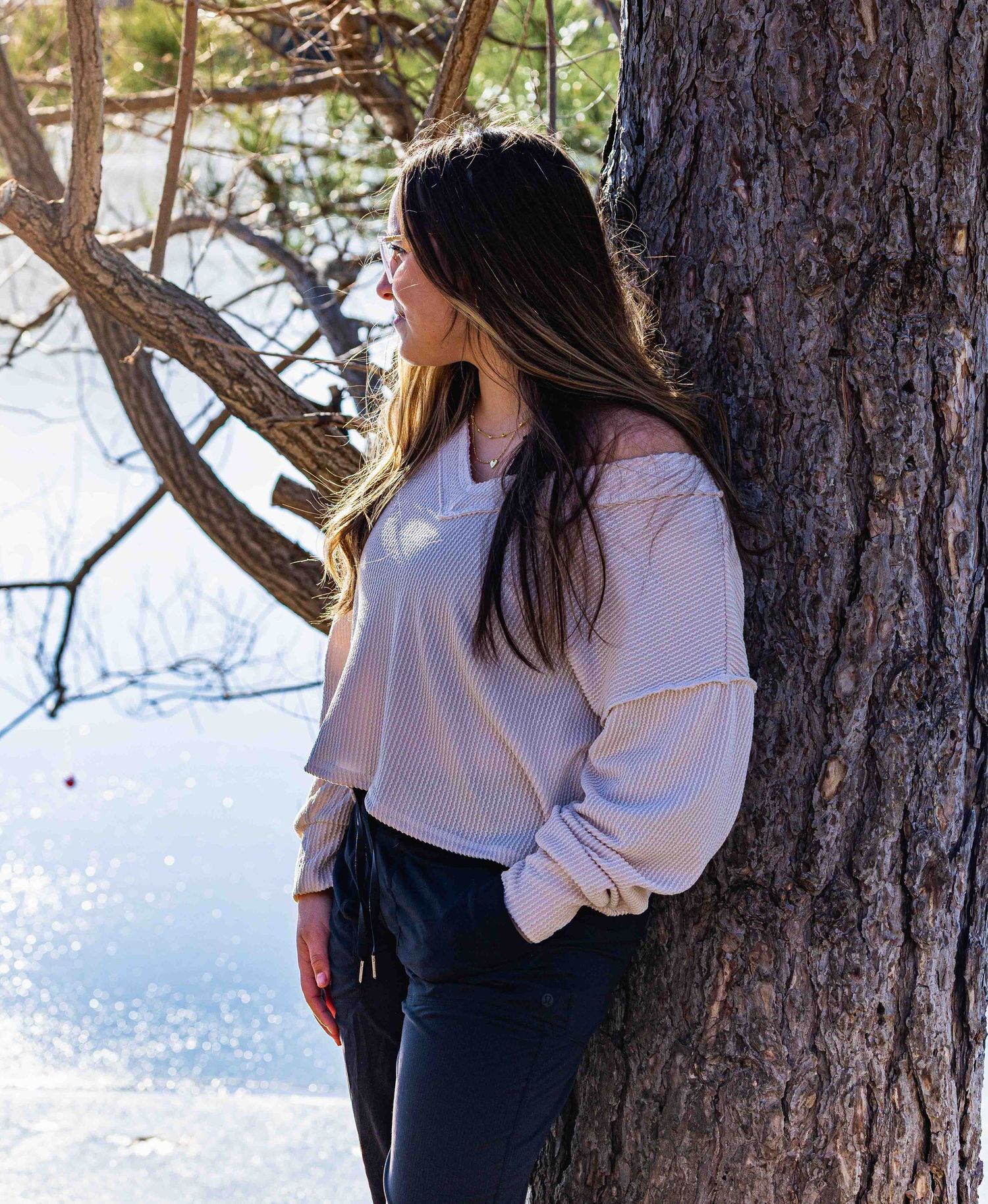 Woman in white off-shoulder top leans against pine tree by a serene lake, long dark hair flowing.