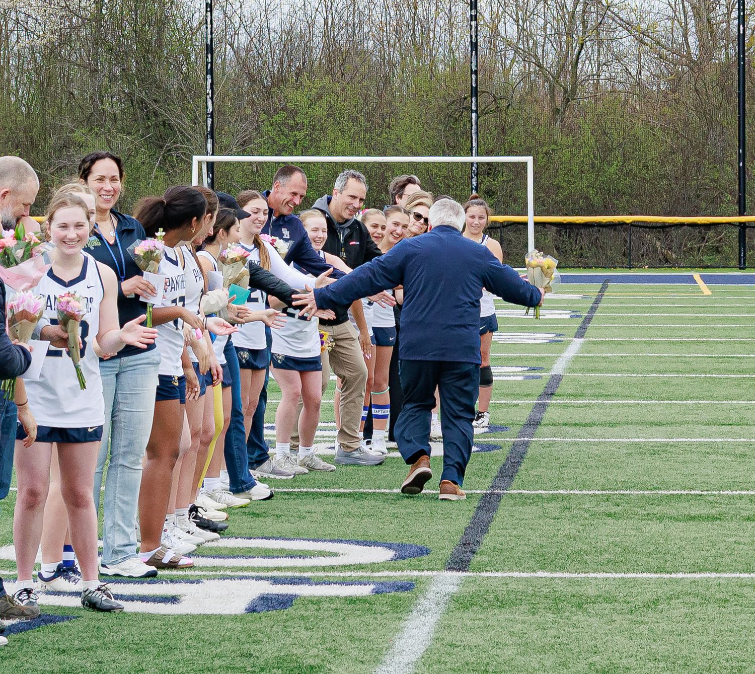 Girls lacrosse team senior day ceremony on turf field, players receiving handshakes from coaches with families present.