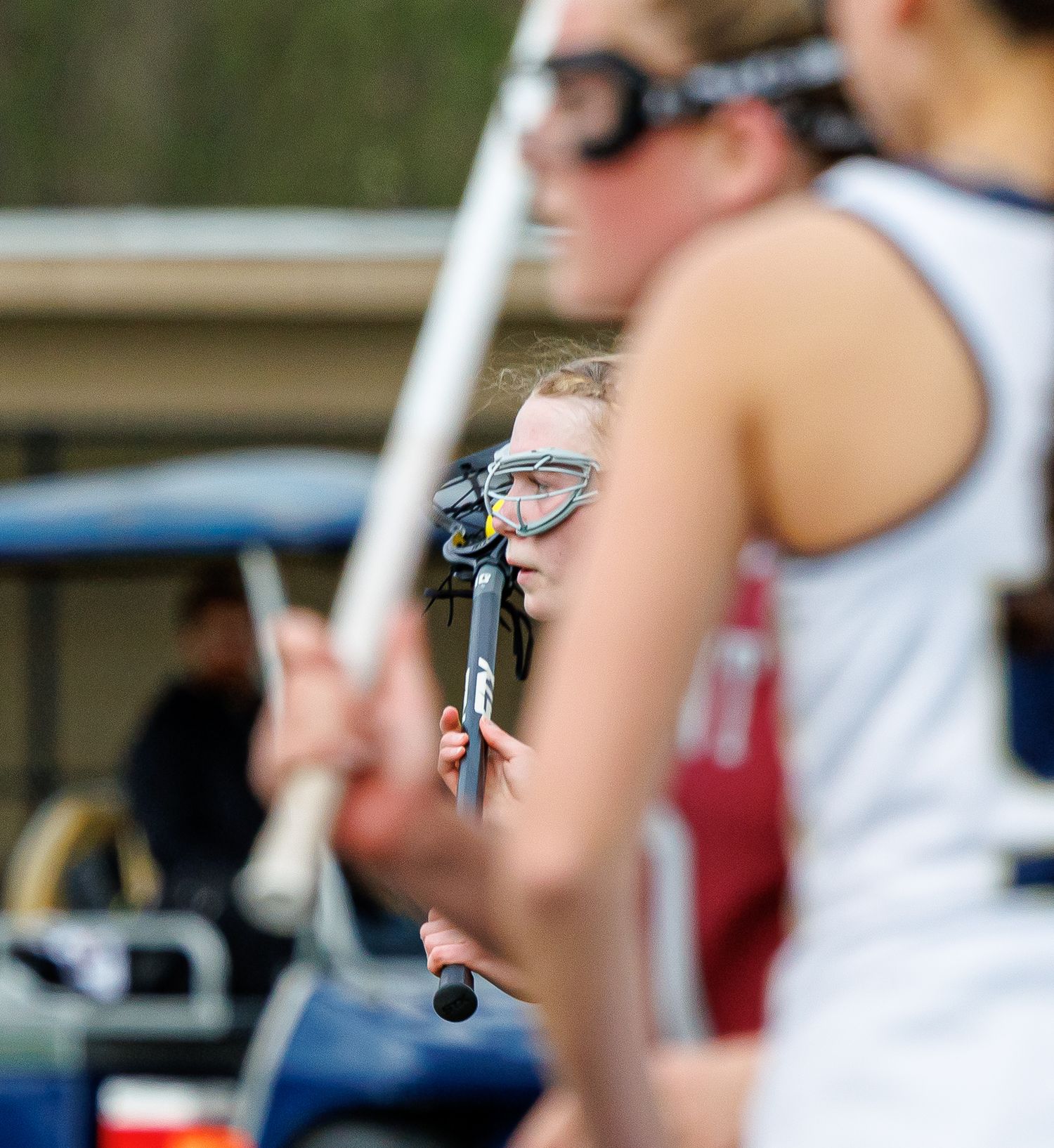 A female lacrosse player peers over her stick wearing protective goggles during a competitive lacrosse match.