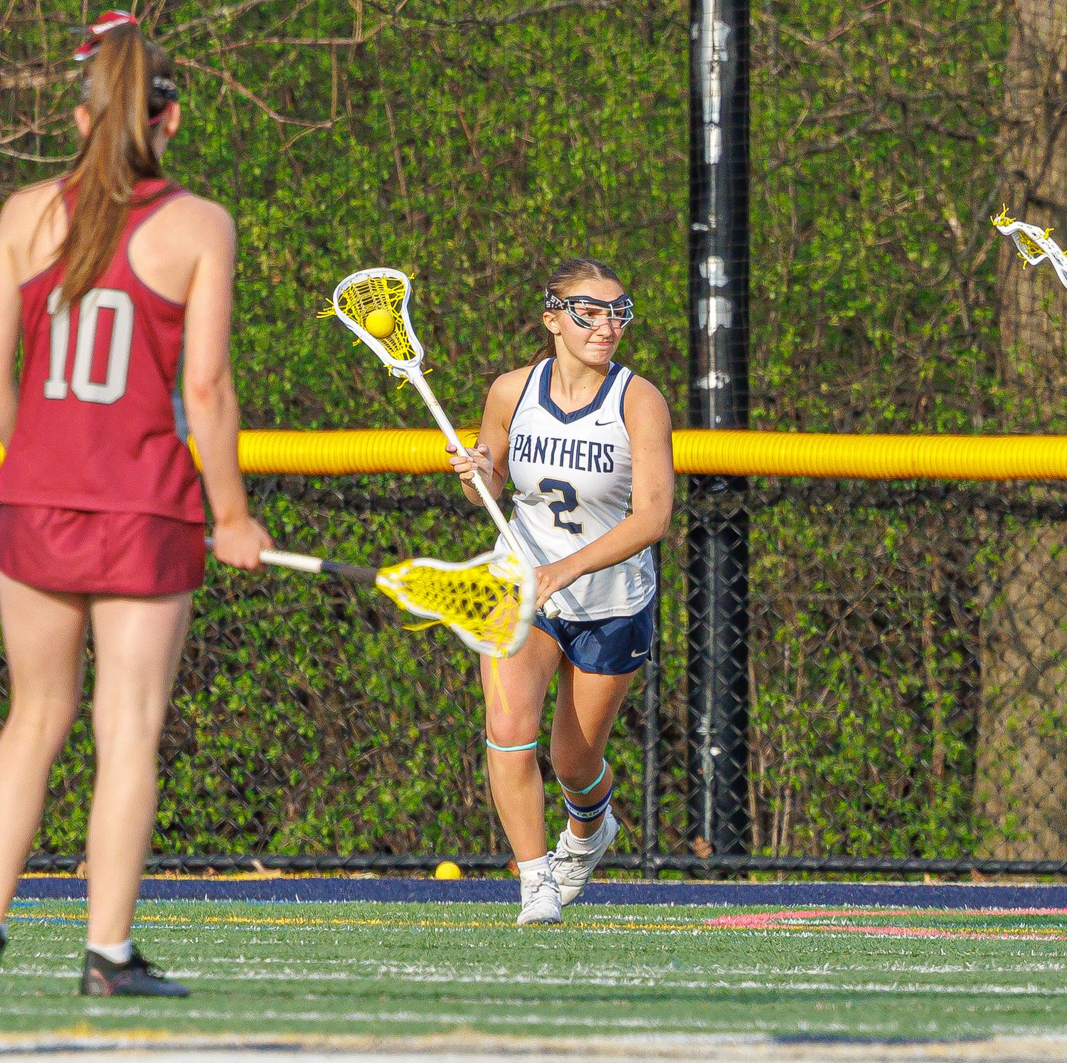 Two female lacrosse players compete on field, Panthers player #2 in white uniform running with stick toward opponent #10.