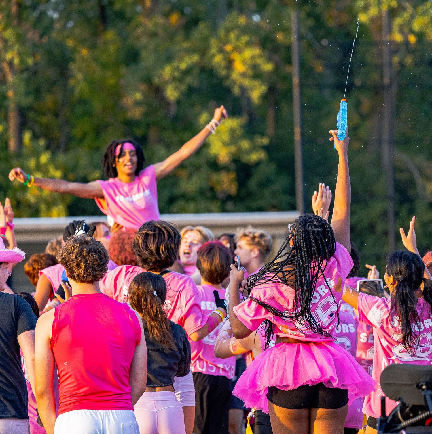Students in pink shirts celebrate outdoors at a school spirit event with raised arms and colorful powder.