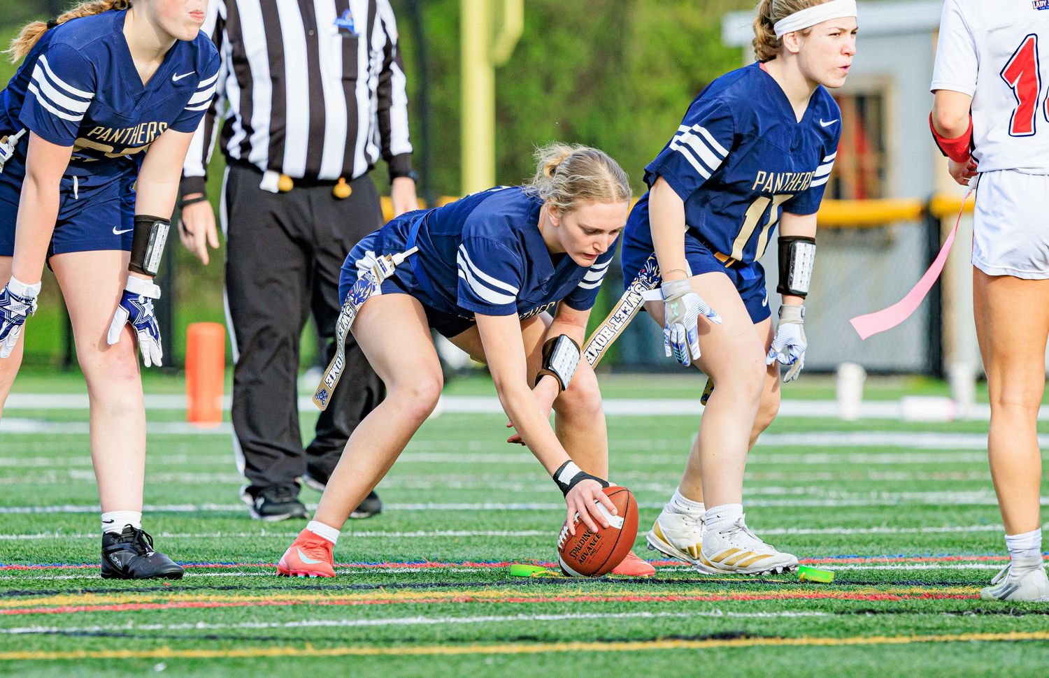 Female flag football player bends to snap football at line of scrimmage during game.