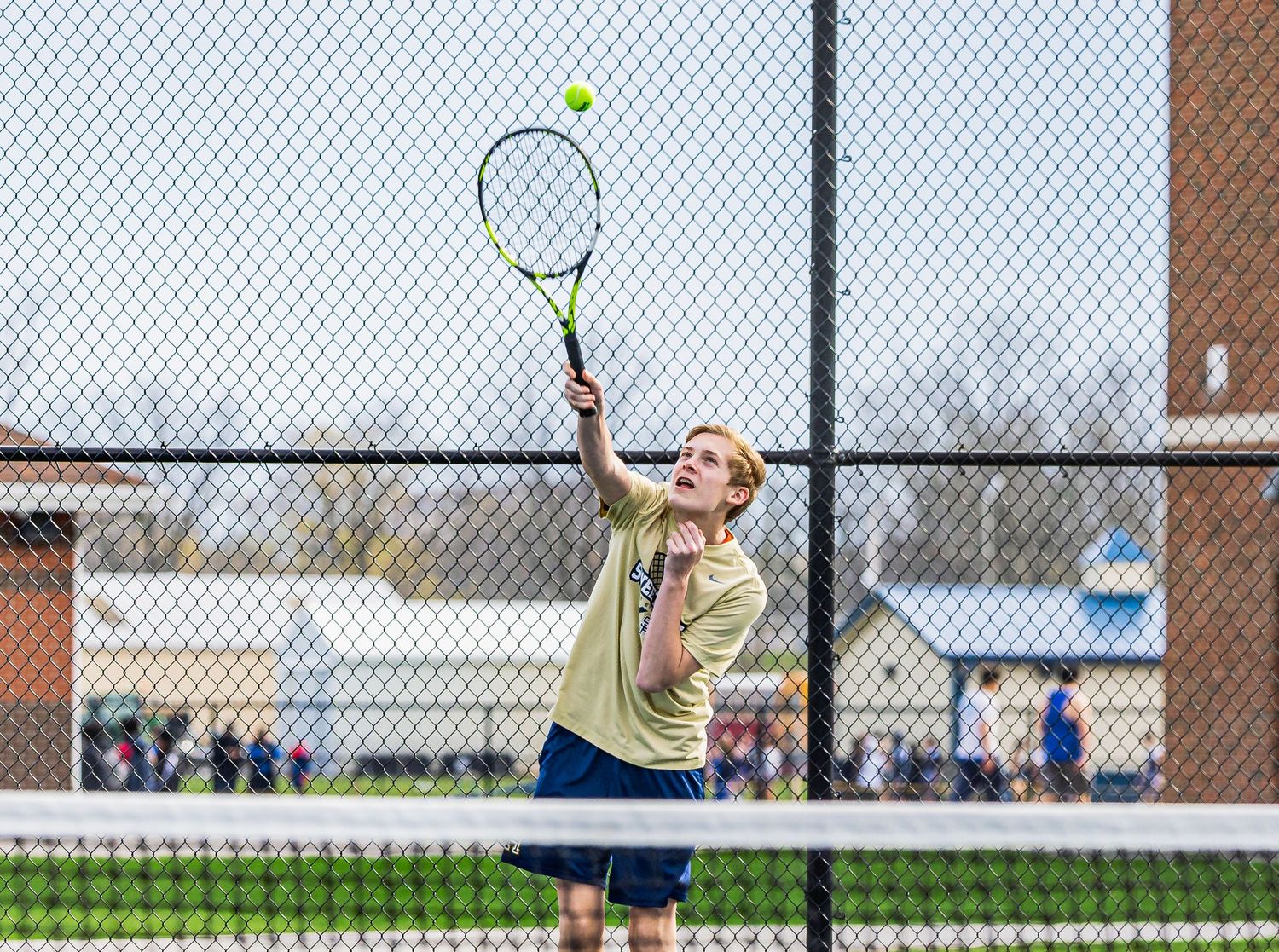 Young tennis player serving ball on outdoor court during match, wearing yellow shirt, reaching racket overhead.