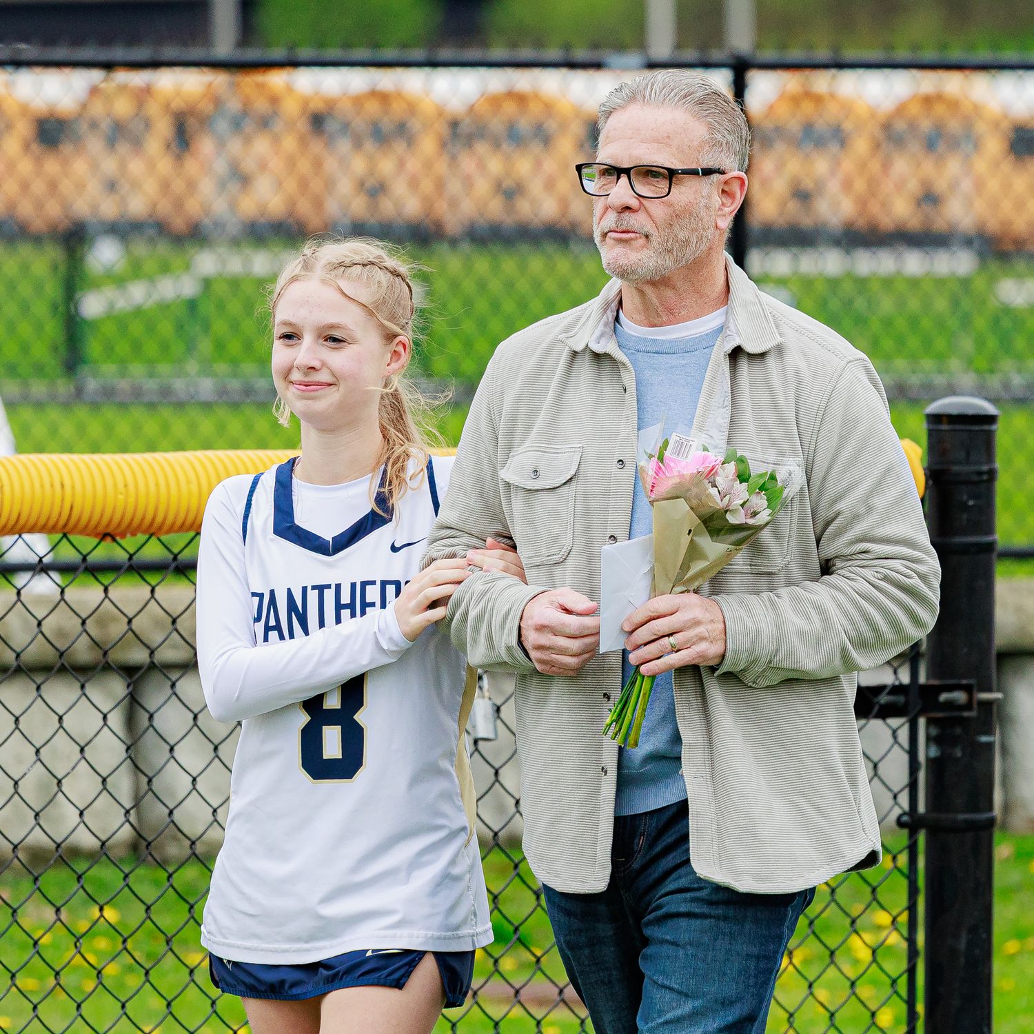 A young female lacrosse player wearing number 8 walks with an older man holding pink flowers at a sports event.