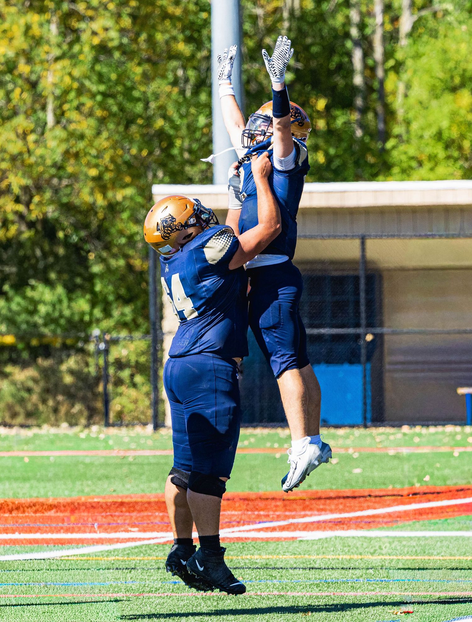 Two football players in blue uniforms celebrate on a green field, one lifting the other in triumph.