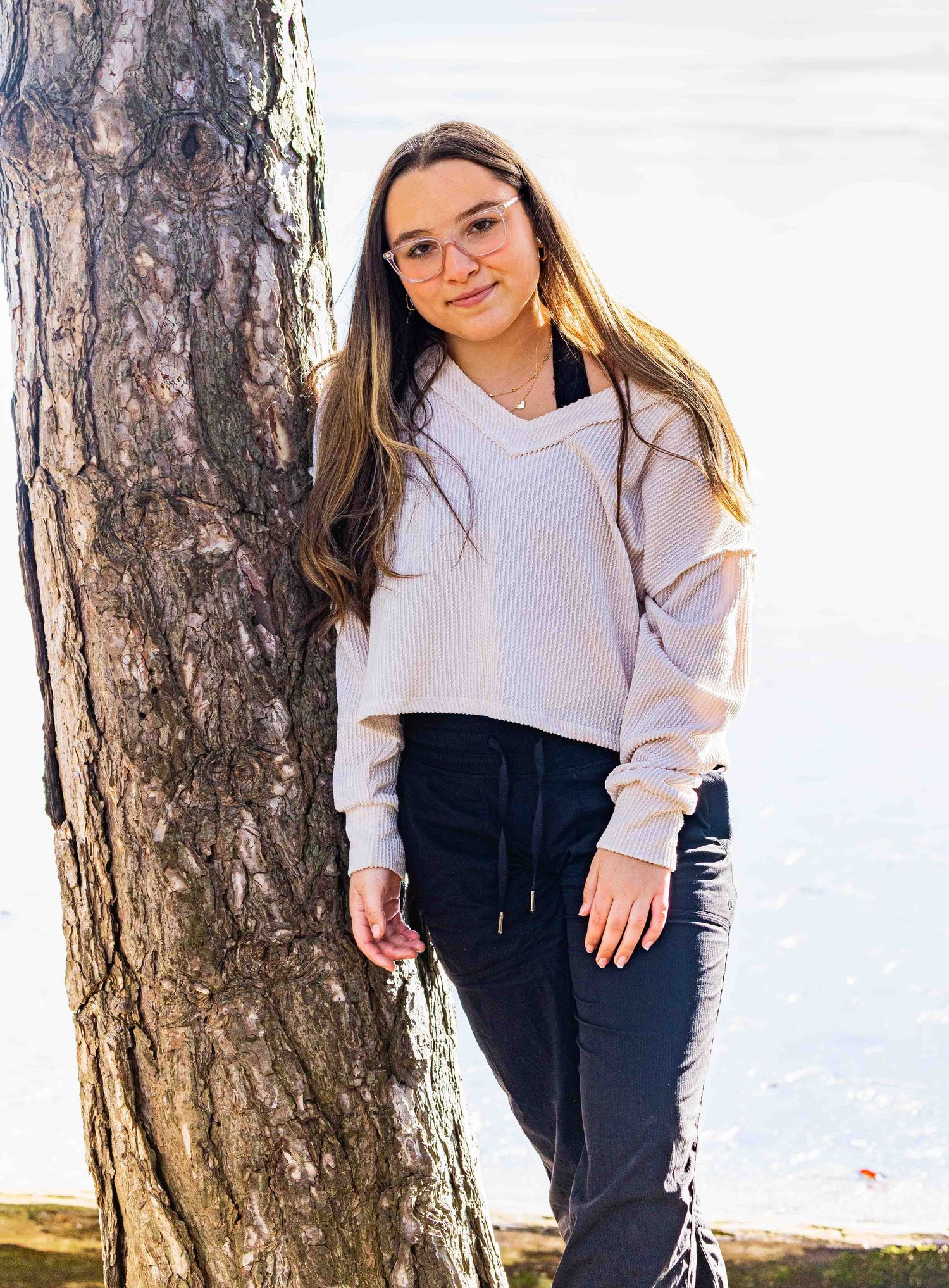 Young woman with long hair and glasses leans against a tree outdoors near water in a light hoodie.