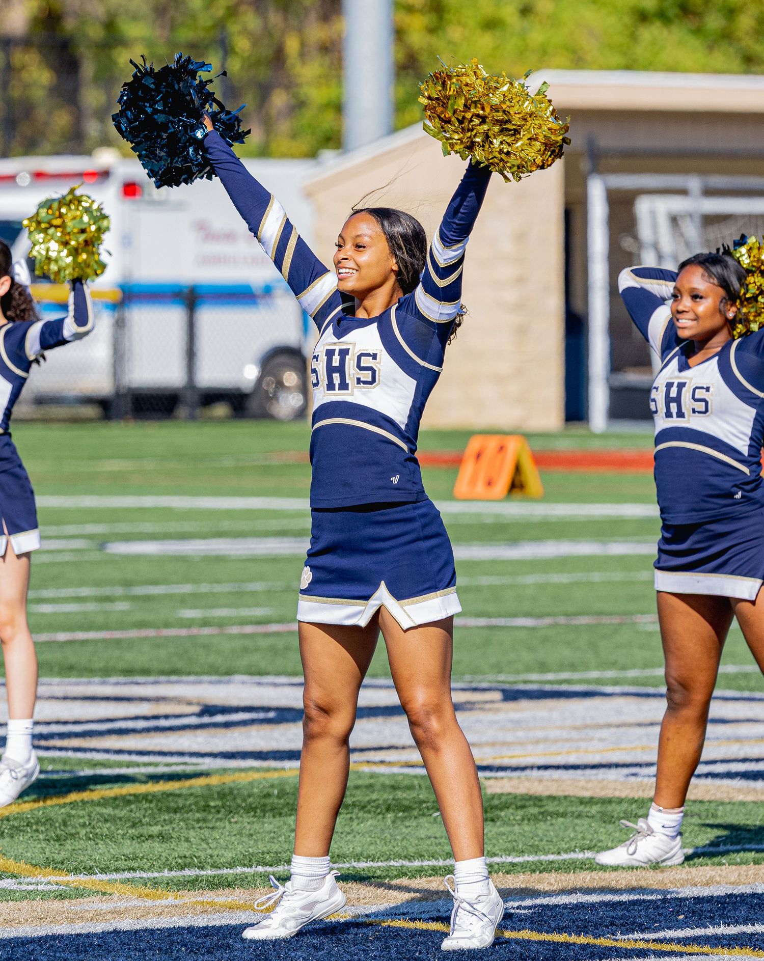 Cheerleader smiling with gold pom-poms raised in navy uniform on football field.