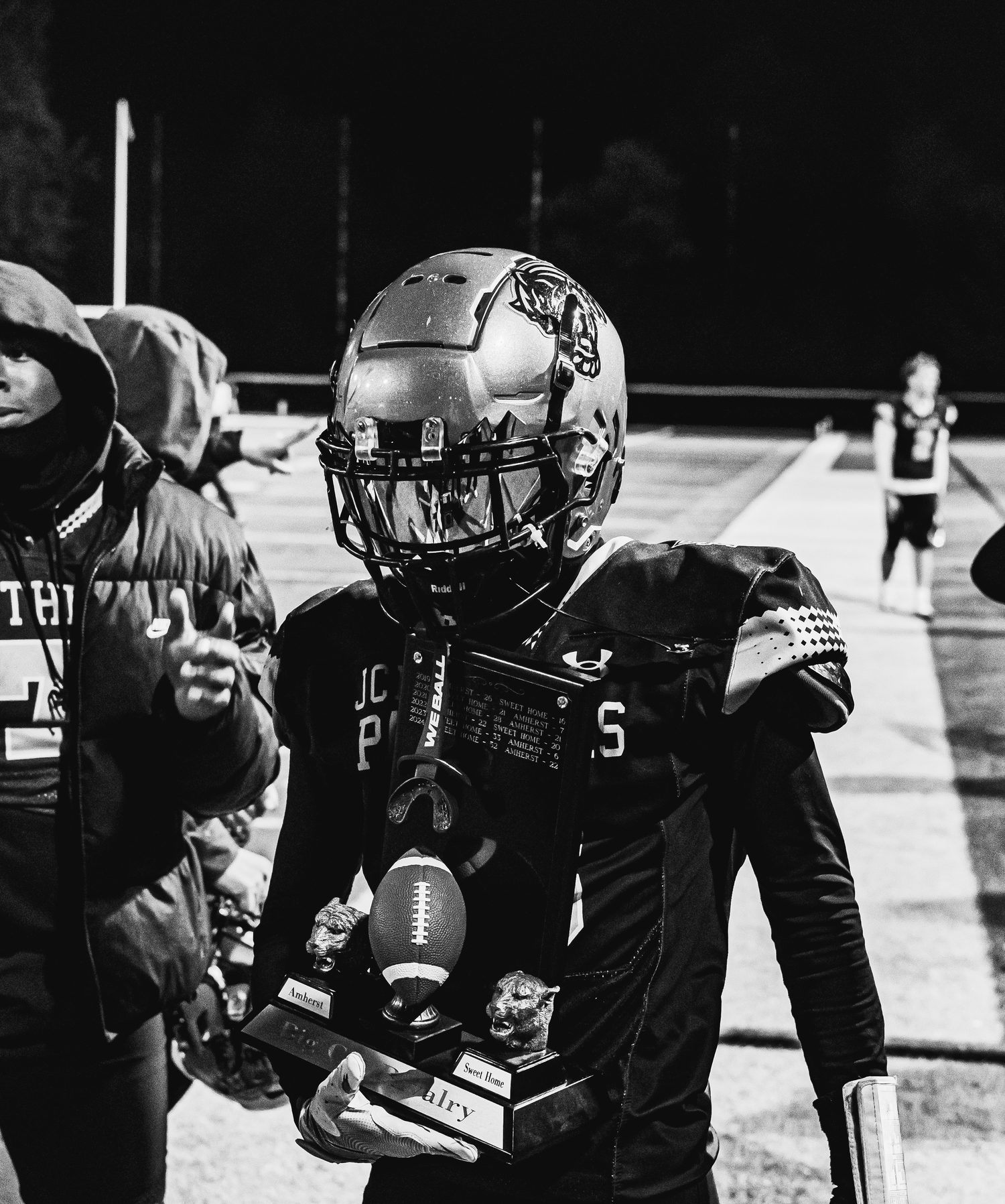 Young football player in helmet and uniform proudly holds a trophy on the field in a black and white photo.