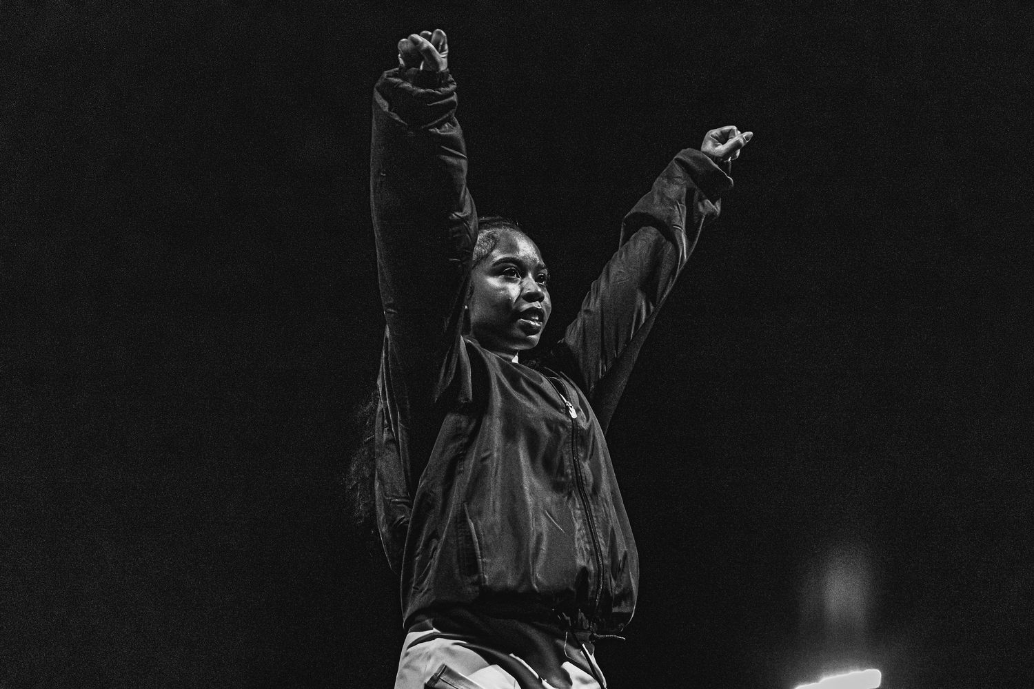 A performer in a dark concert venue raises their arms while singing on stage during an energetic performance.