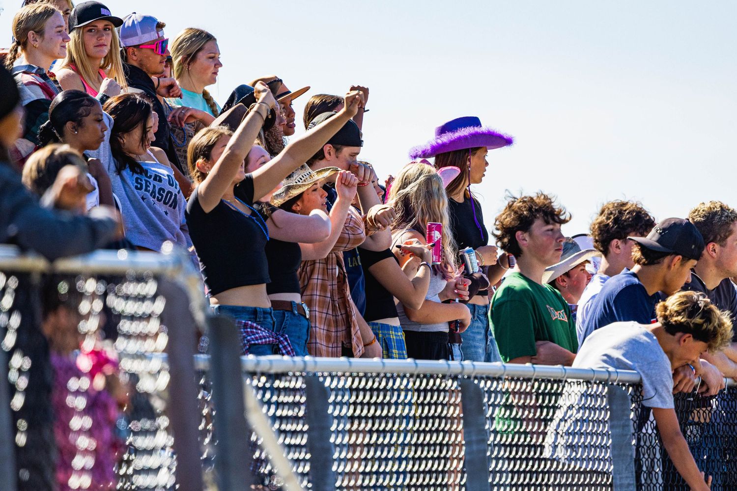 Spectators at an outdoor event take photos and watch from behind a metal railing on a sunny day.