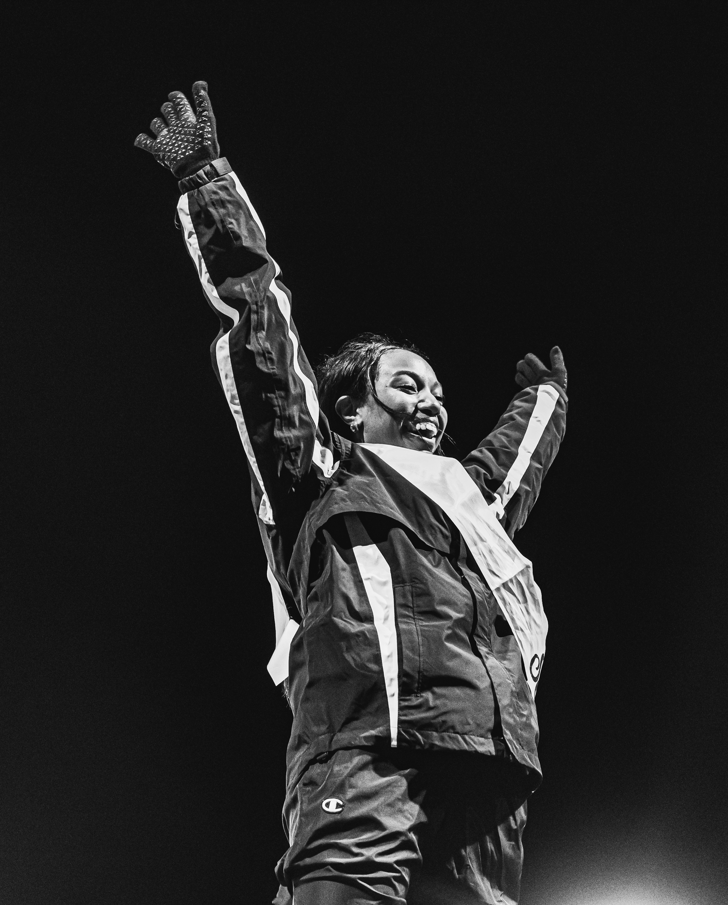 Black and white photo of an athlete in a Champion jacket celebrating with arms raised under stadium lights.