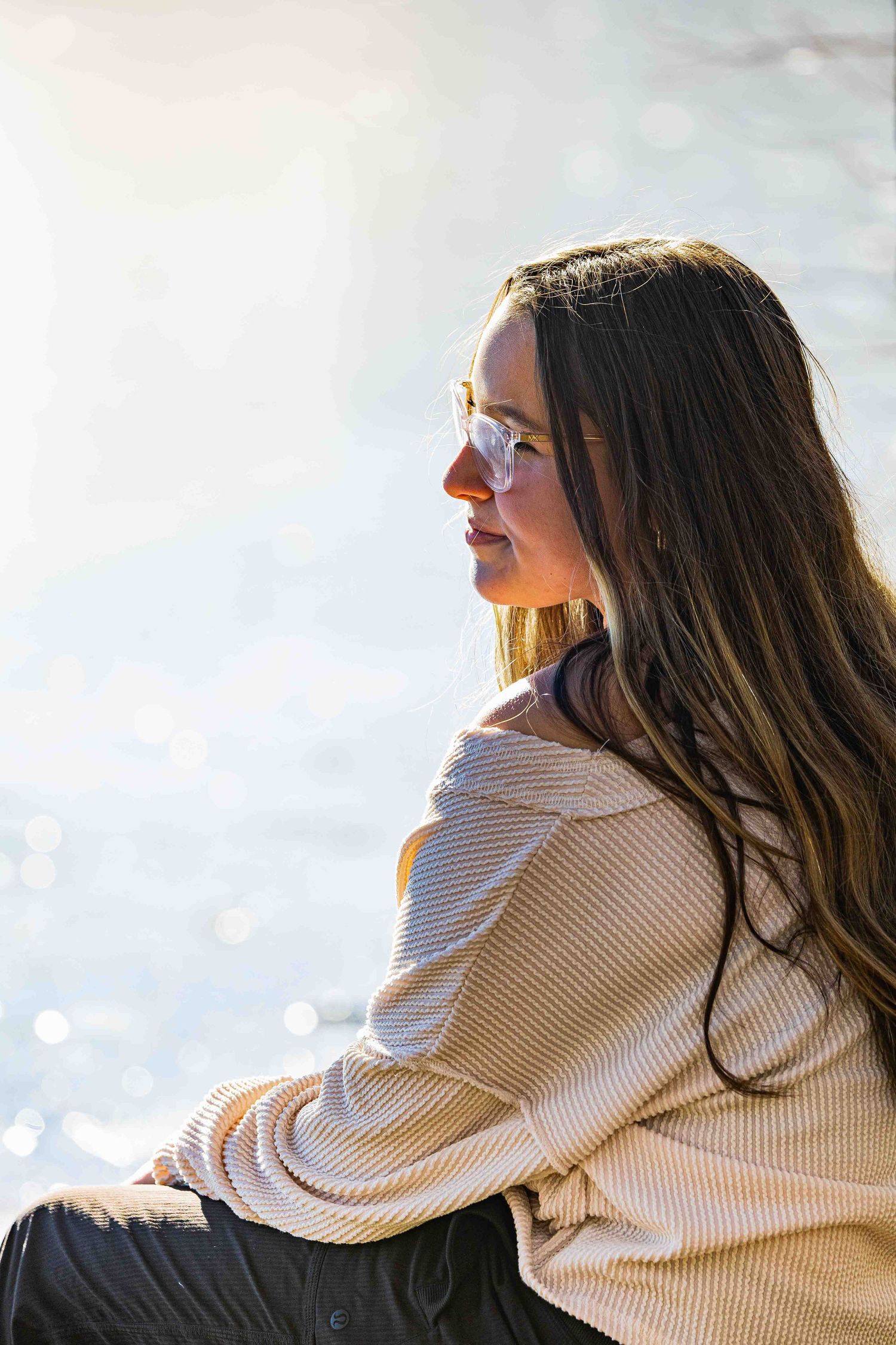 Young woman with long dark hair and glasses sits outdoors by shimmering water in golden sunlight wearing a striped top.
