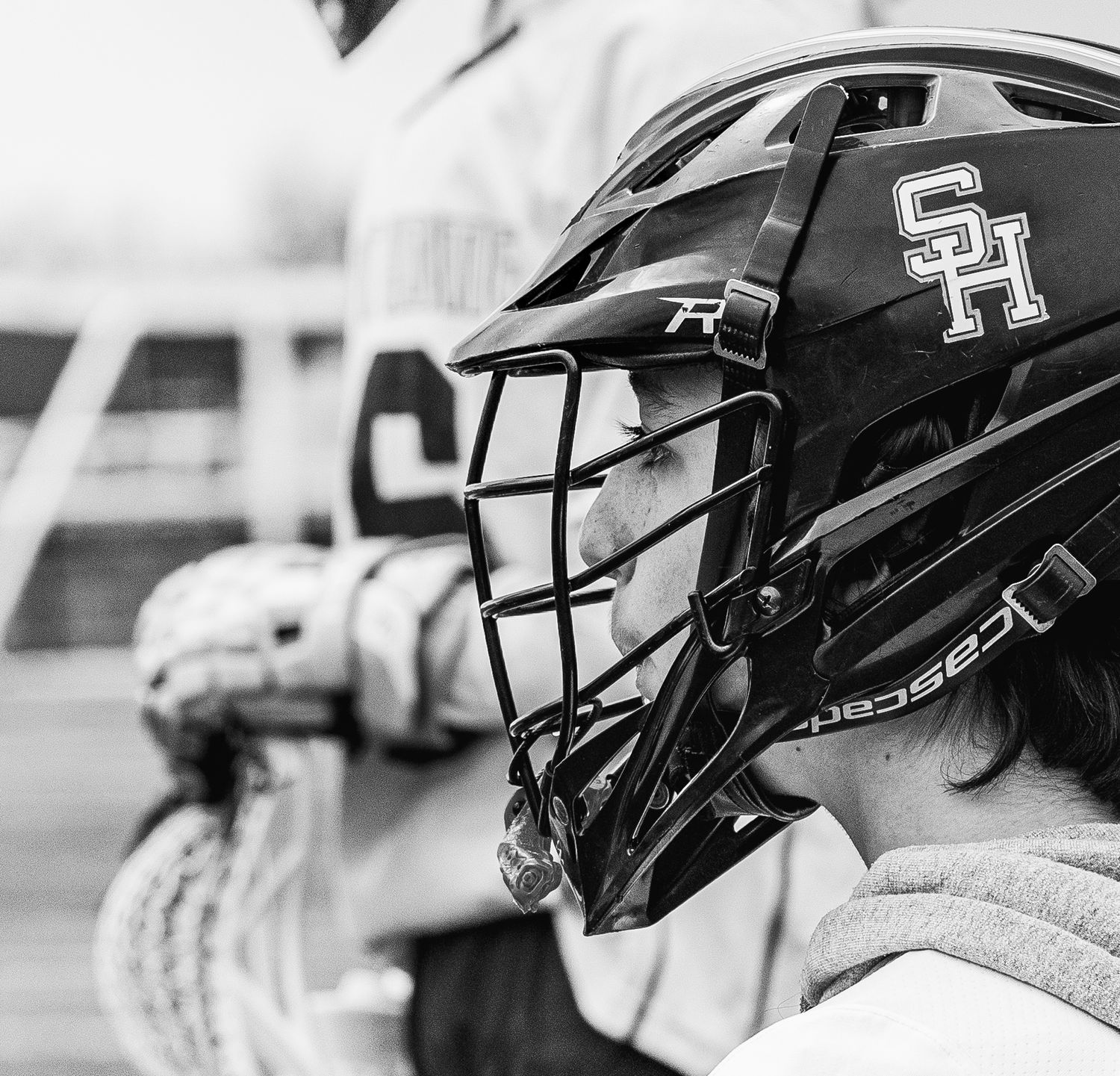 Black and white photo of a lacrosse player wearing an SH helmet with cage face guard holding a lacrosse stick.