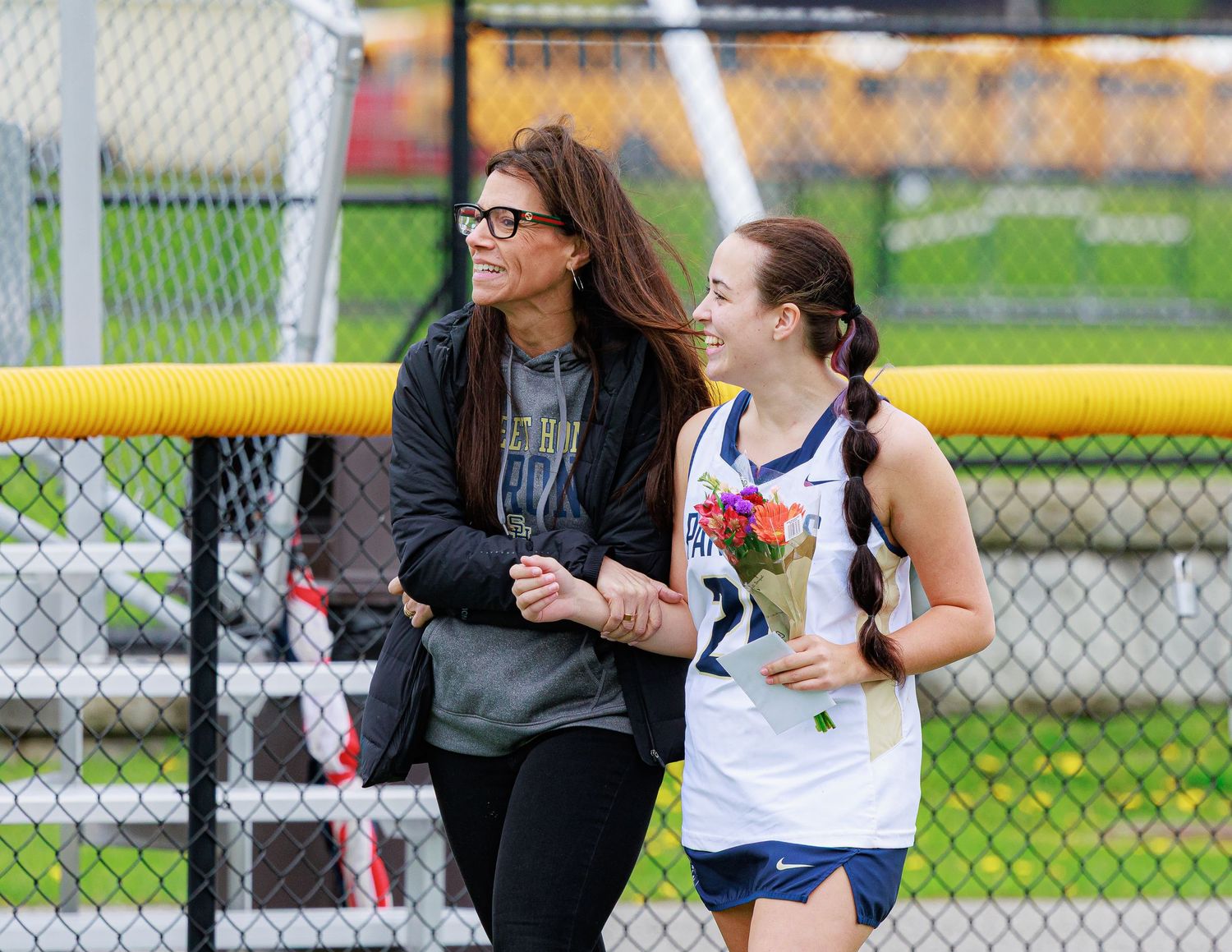A female athlete in uniform holds flowers while walking with a woman in black jacket at a track and field event.