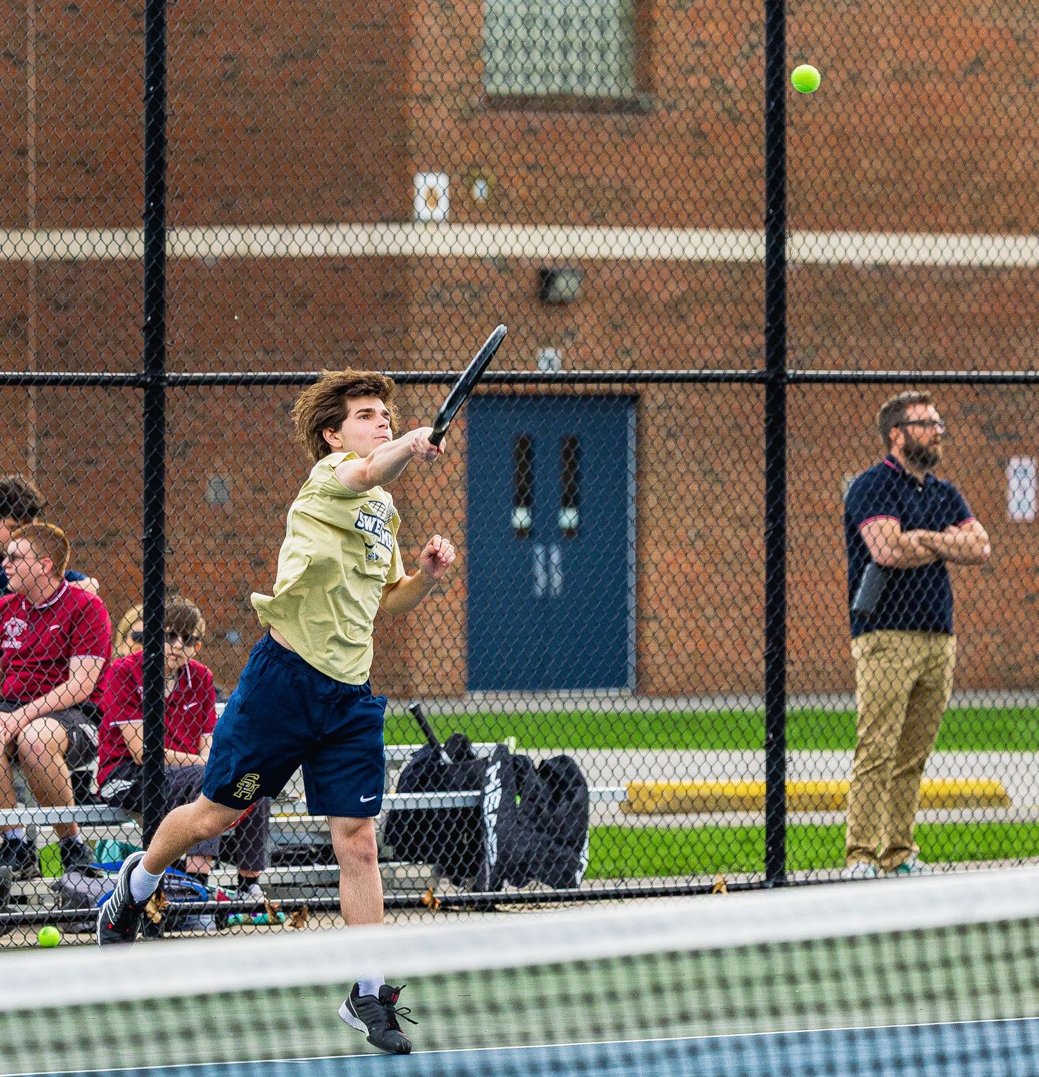 Young tennis player mid-swing on outdoor court, with coach and spectators watching near chain-link fence.