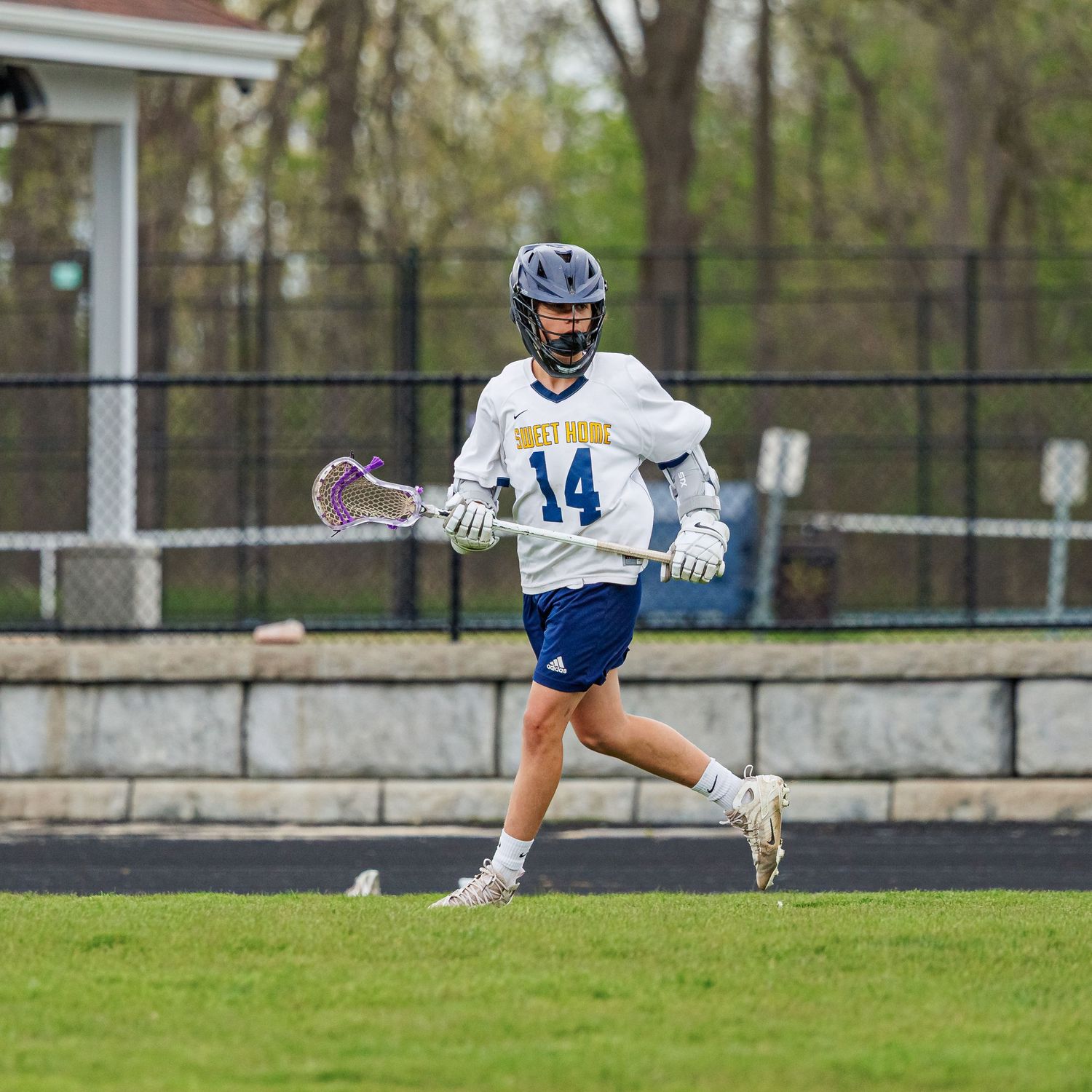 Lacrosse player #14 in white uniform runs with stick on outdoor field during spring game.