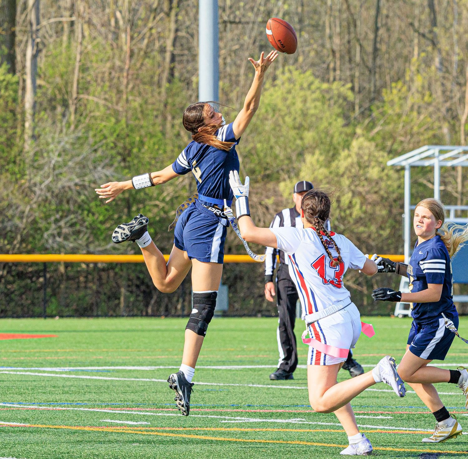 Female flag football player leaps high to catch football over defending opponent.