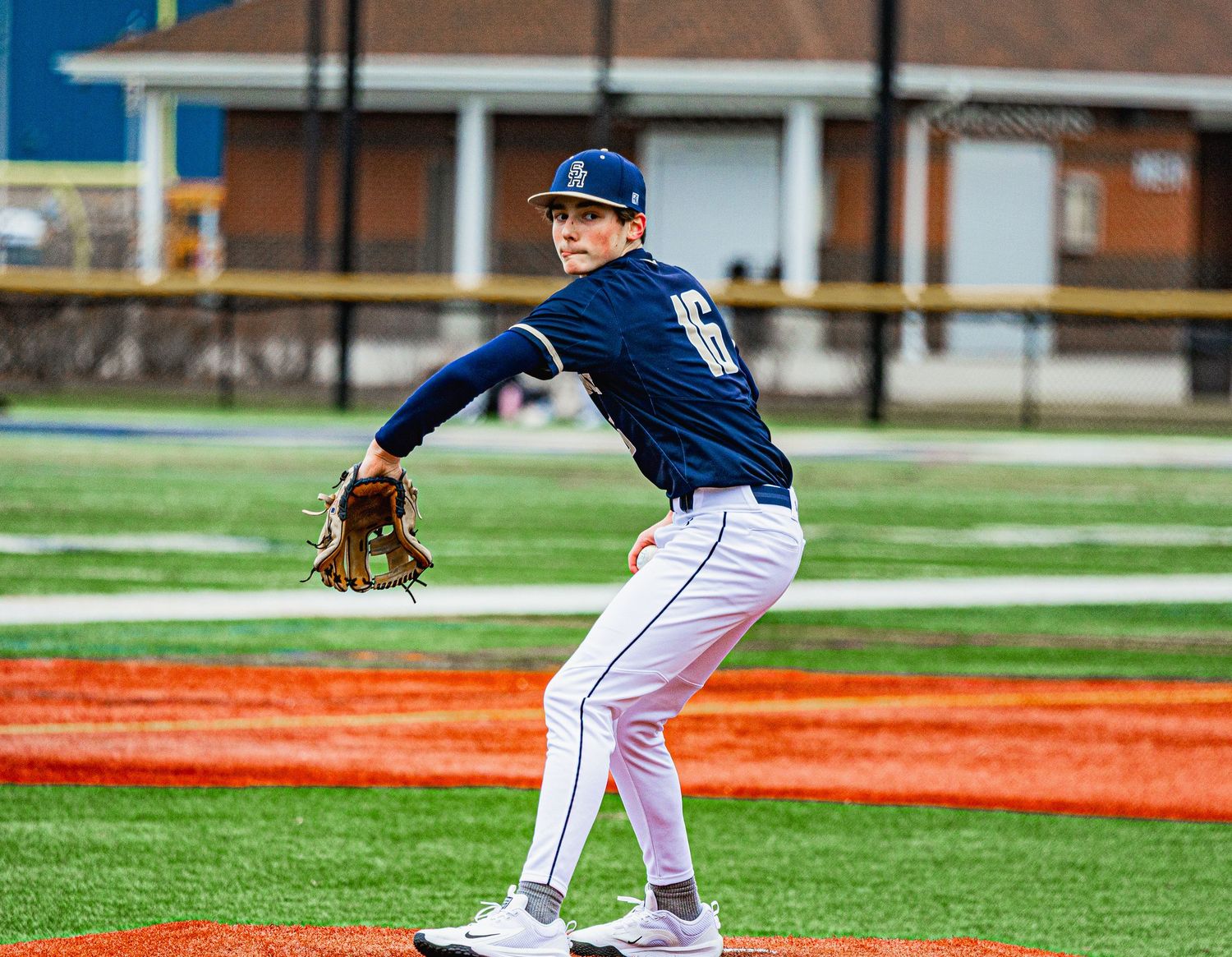 Baseball pitcher in navy uniform winds up to throw on colorful artificial turf field.