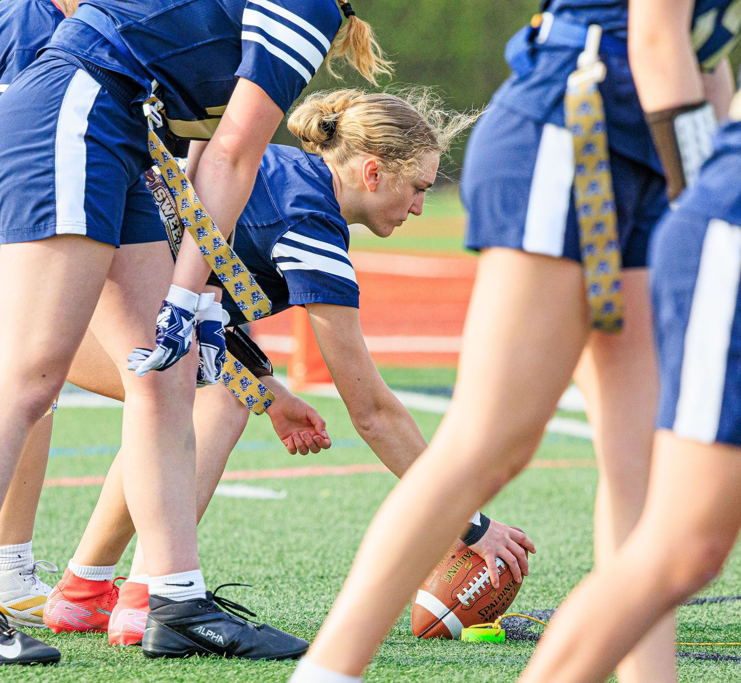 Female flag football player crouches over football at line of scrimmage on turf field.