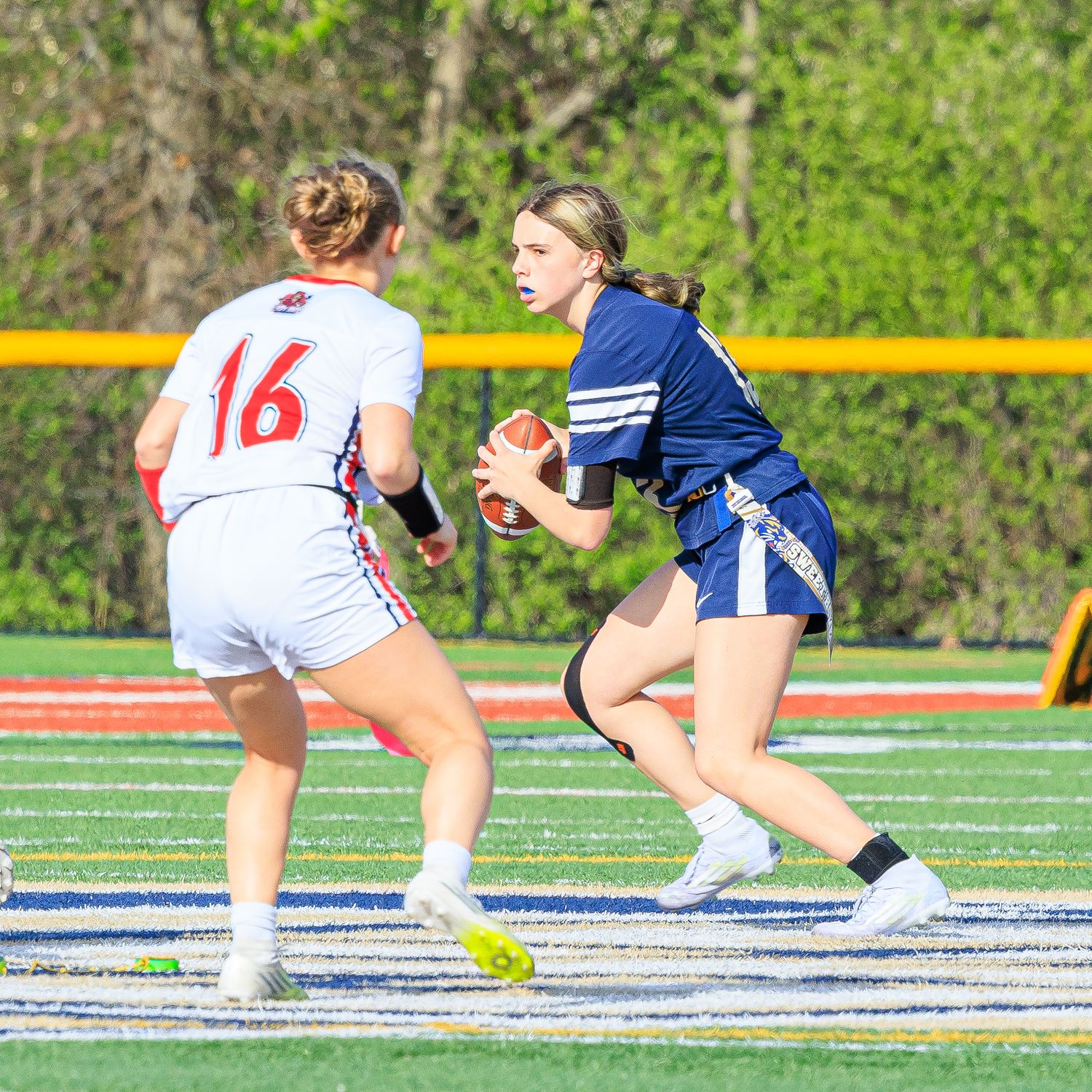 Female flag football player in blue jersey holds ball while evading defender wearing number 16 white jersey on turf field.