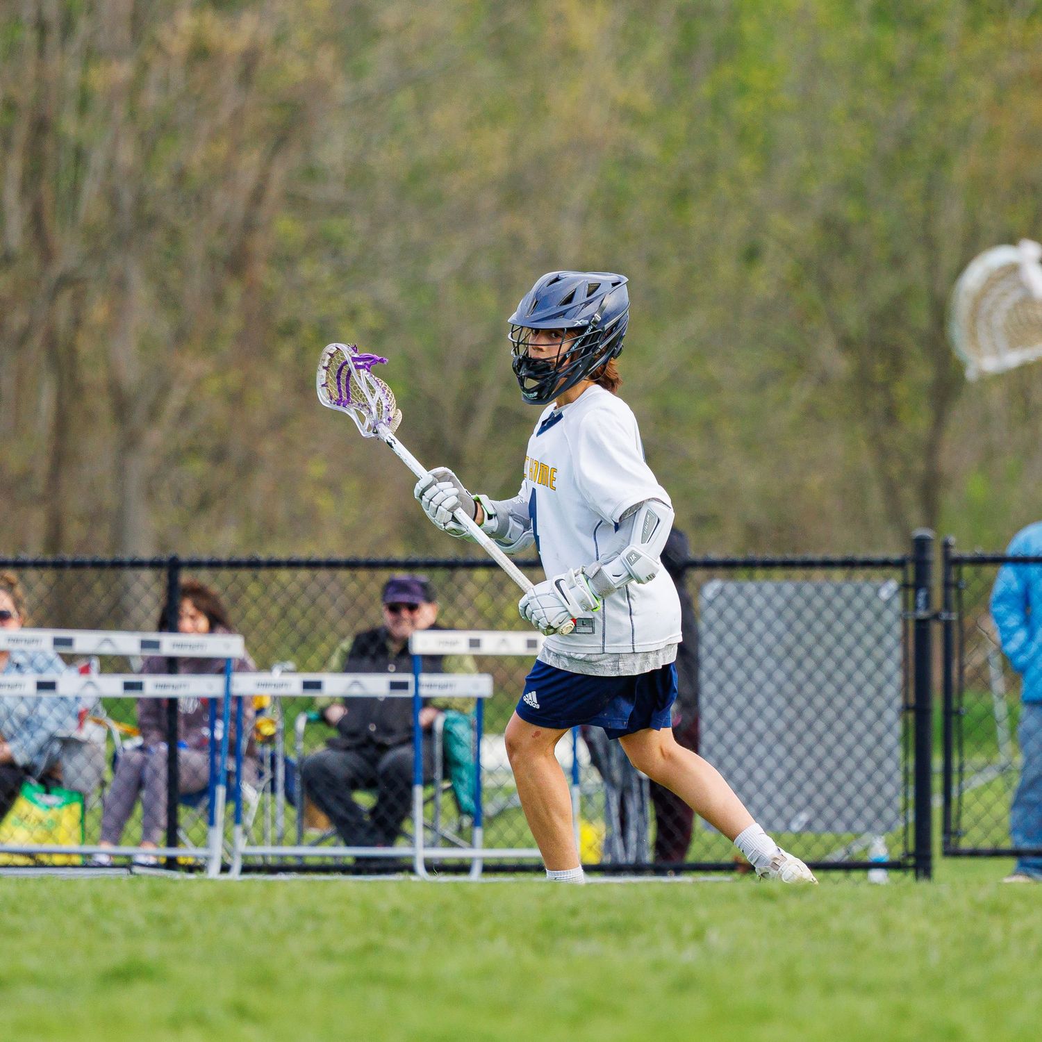 A lacrosse player in navy and gold uniform runs with stick on a green field during a spring game.