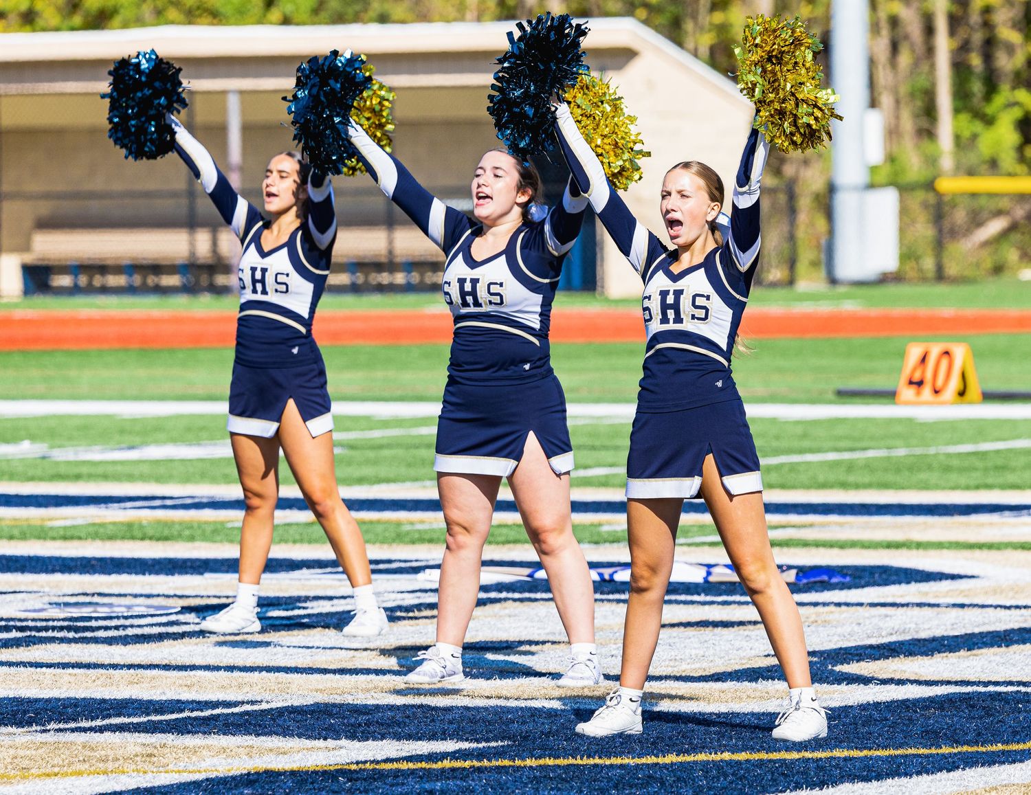 Three high school cheerleaders in navy uniforms perform energetically on a football field, waving blue and gold pom-poms.