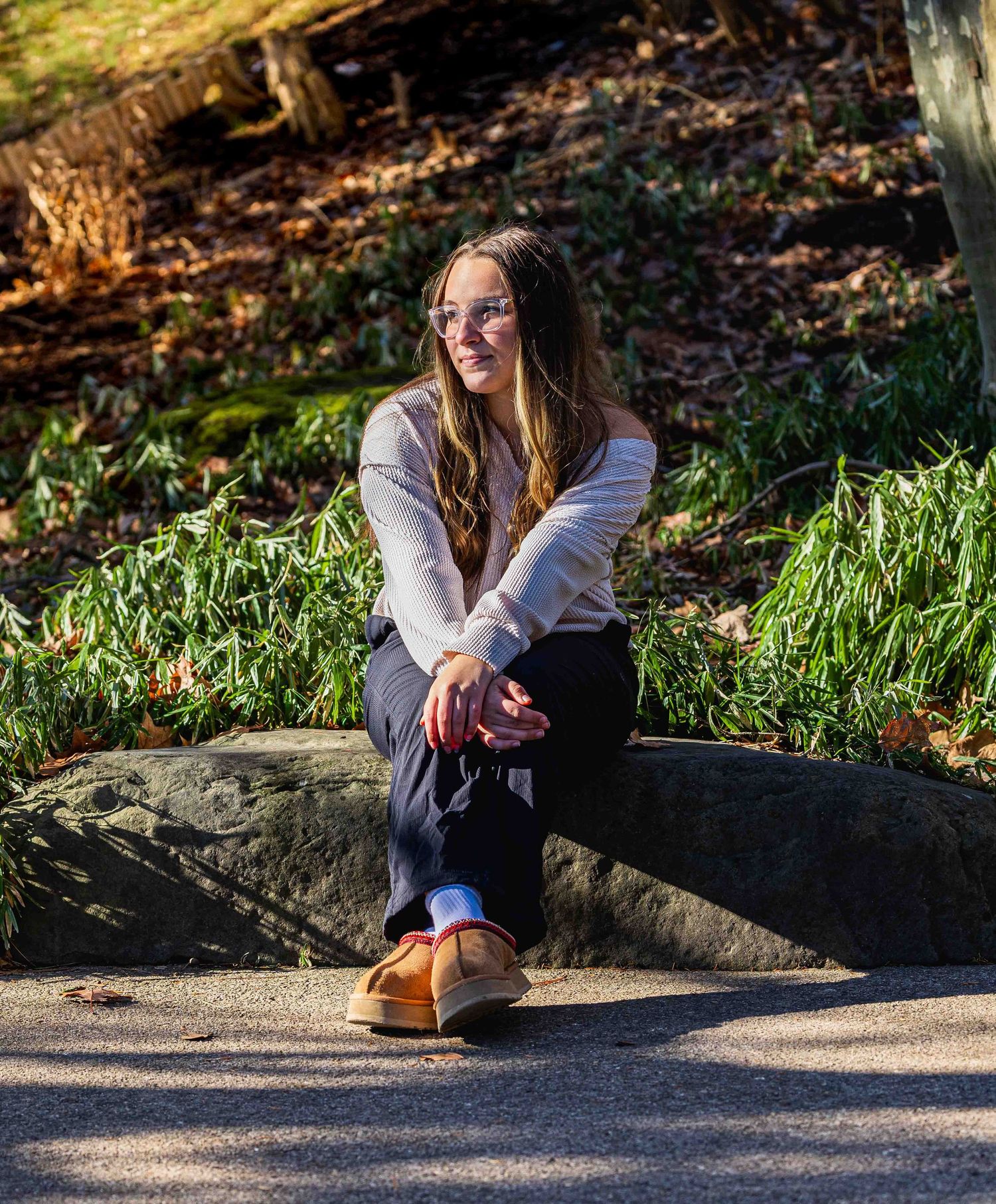 Person in casual attire sitting on a stone ledge in a park setting during golden hour sunlight.