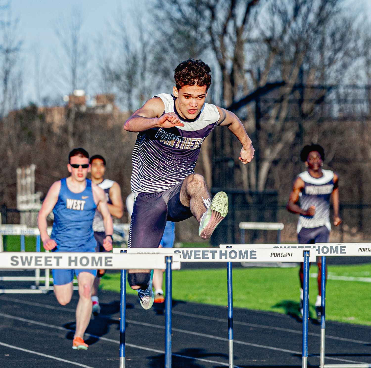 Young athlete clears a hurdle at Sweet Home track meet, with competitors running behind on a sunny day.