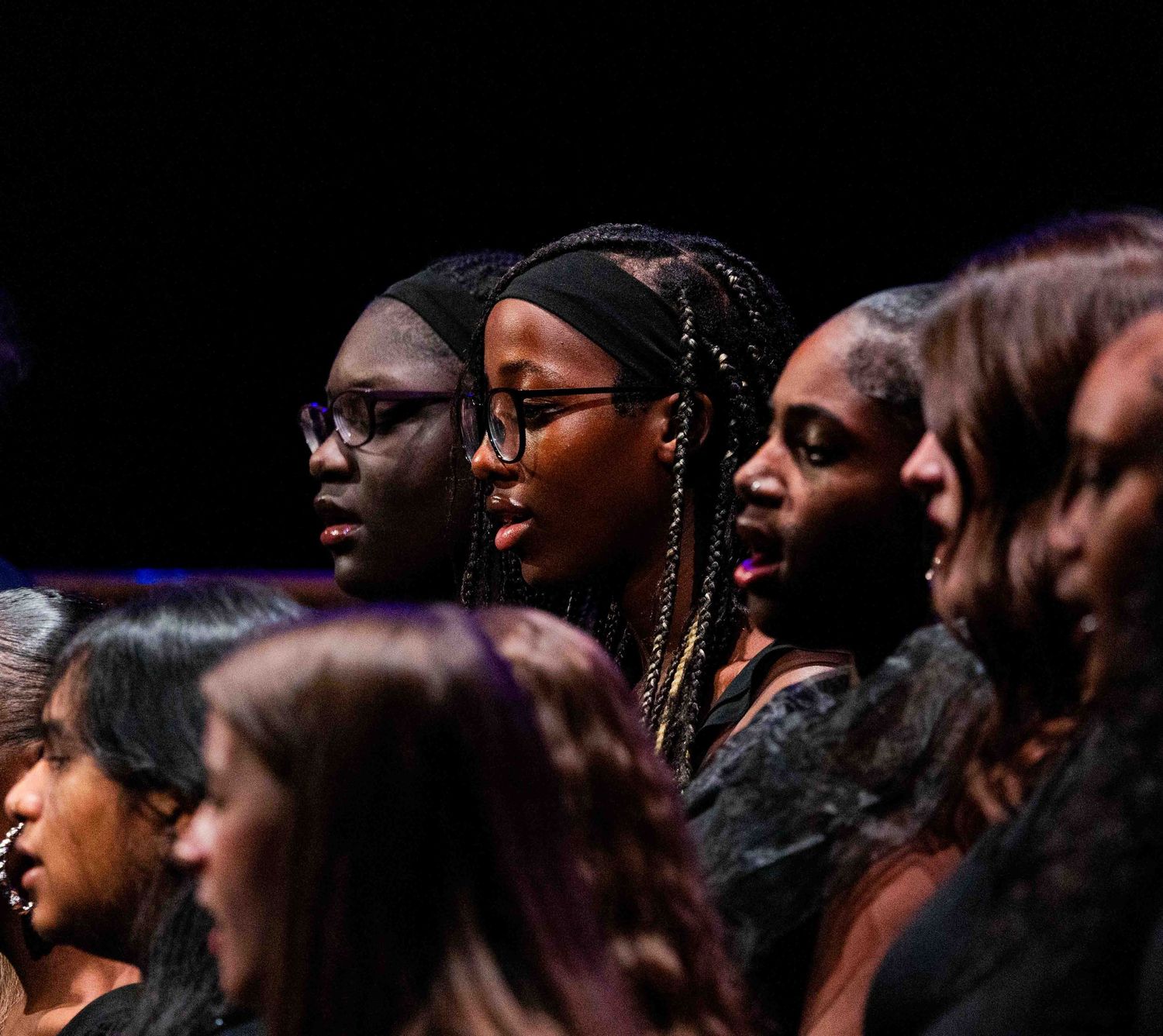 A diverse choir performs on stage in black attire under dramatic lighting during a formal concert event.