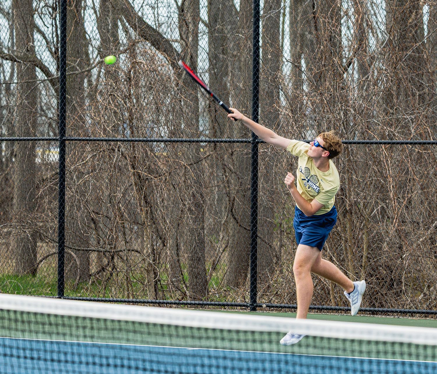 A tennis player leaps to serve, striking a yellow ball with a red racket on an outdoor court surrounded by bare trees.